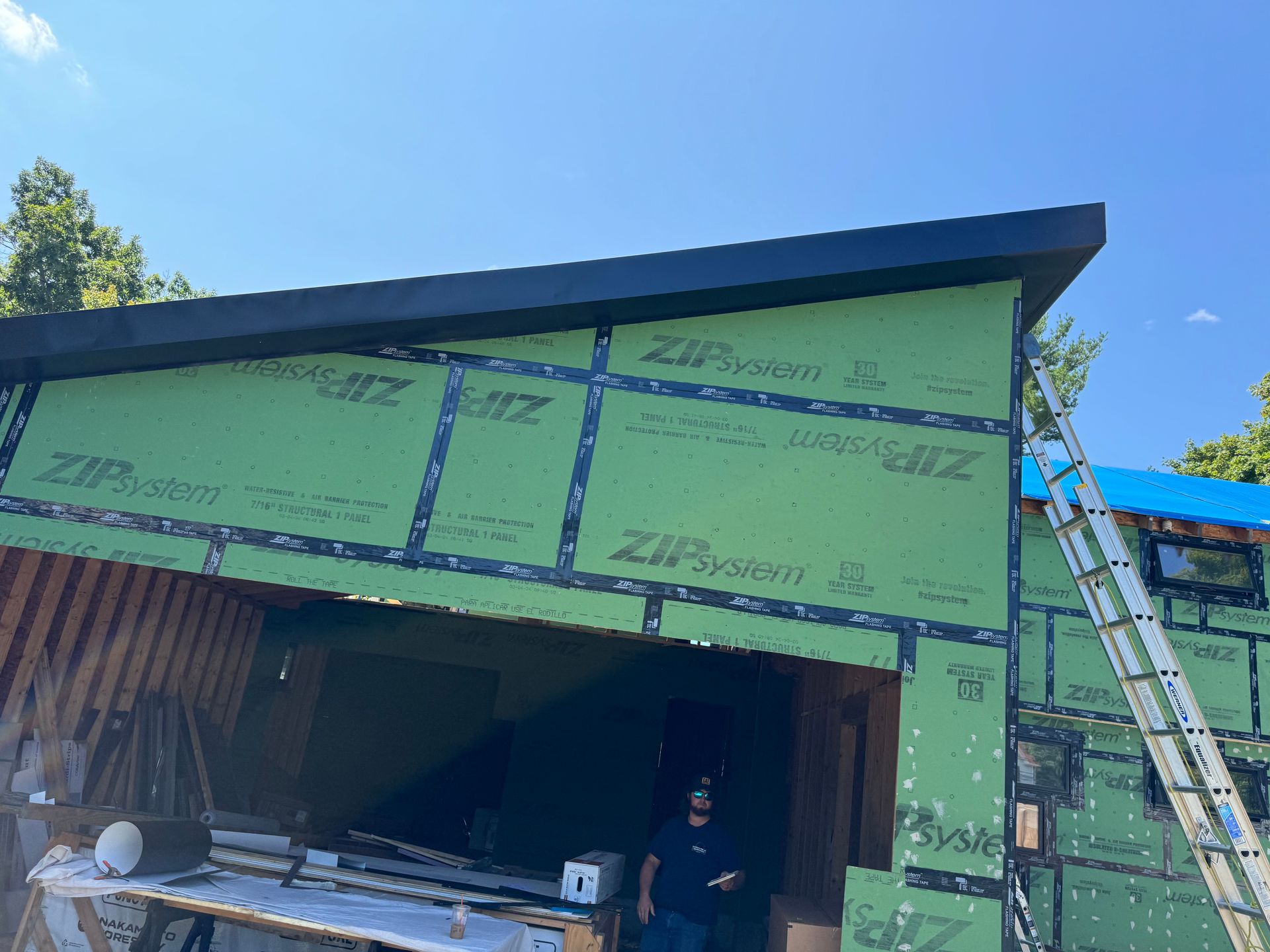 Construction site with green sheathing on a building, person standing, ladder, blue sky.