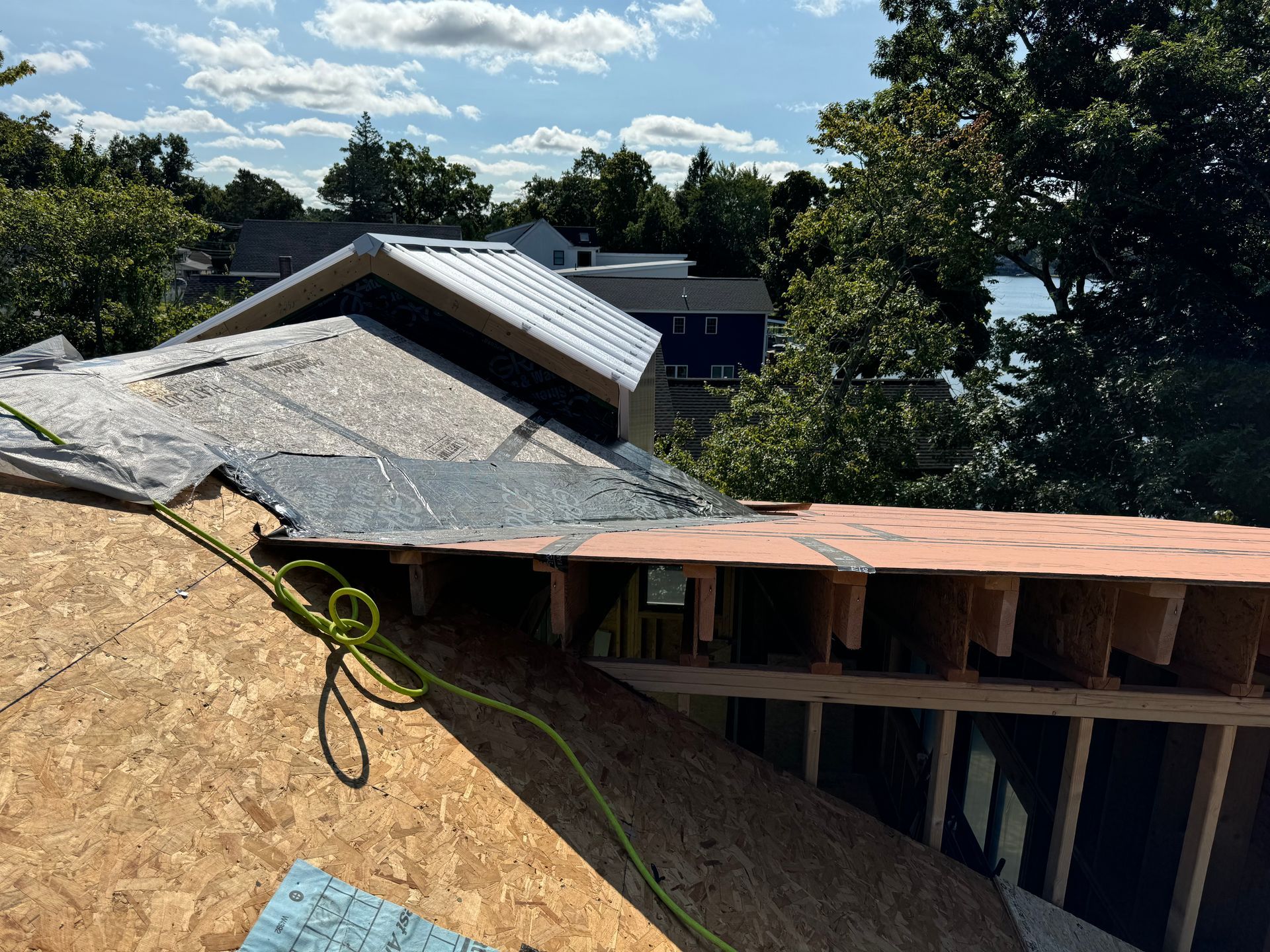 Rooftop construction: plywood, exposed beams, partially shingled. Green rope coiled. Trees and sky in background.