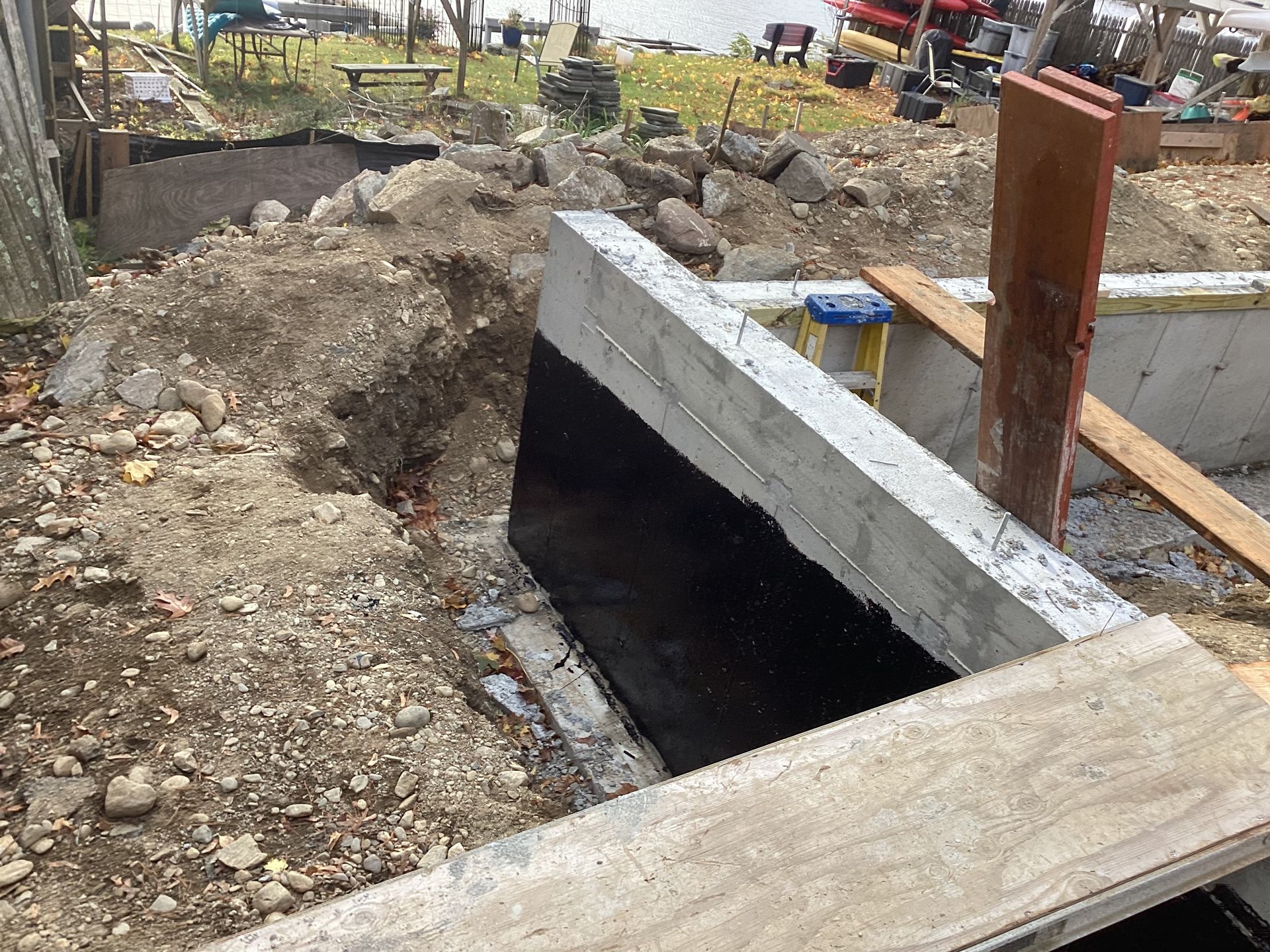 Concrete wall under construction, covered in a black membrane. Dirt, rocks, and wood planks surround it outdoors.