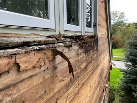 Damaged siding on a house under a window with exposed wood and peeling layers.