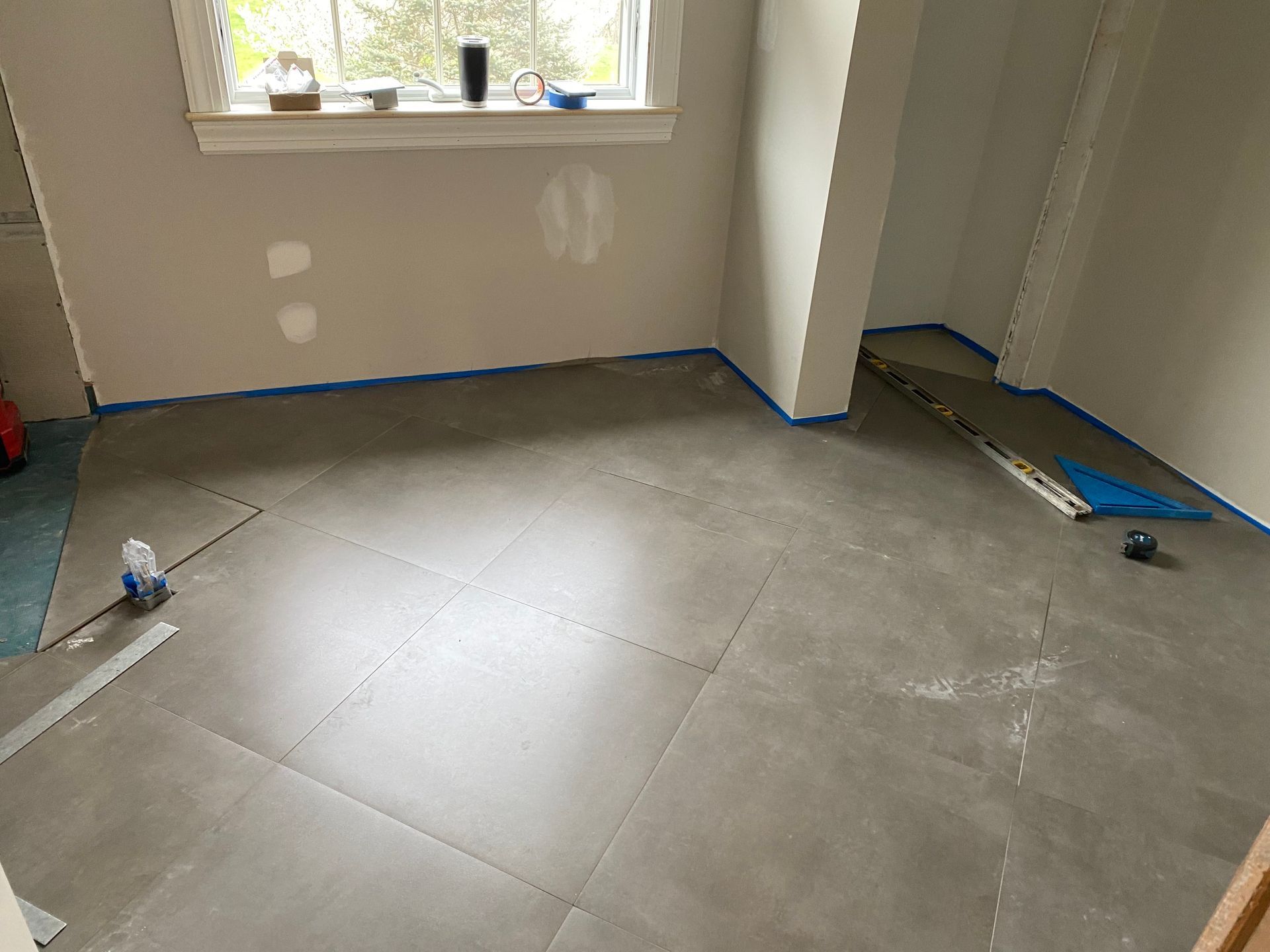 Room with gray tile floor installation in progress, blue tape along edges, window at top.
