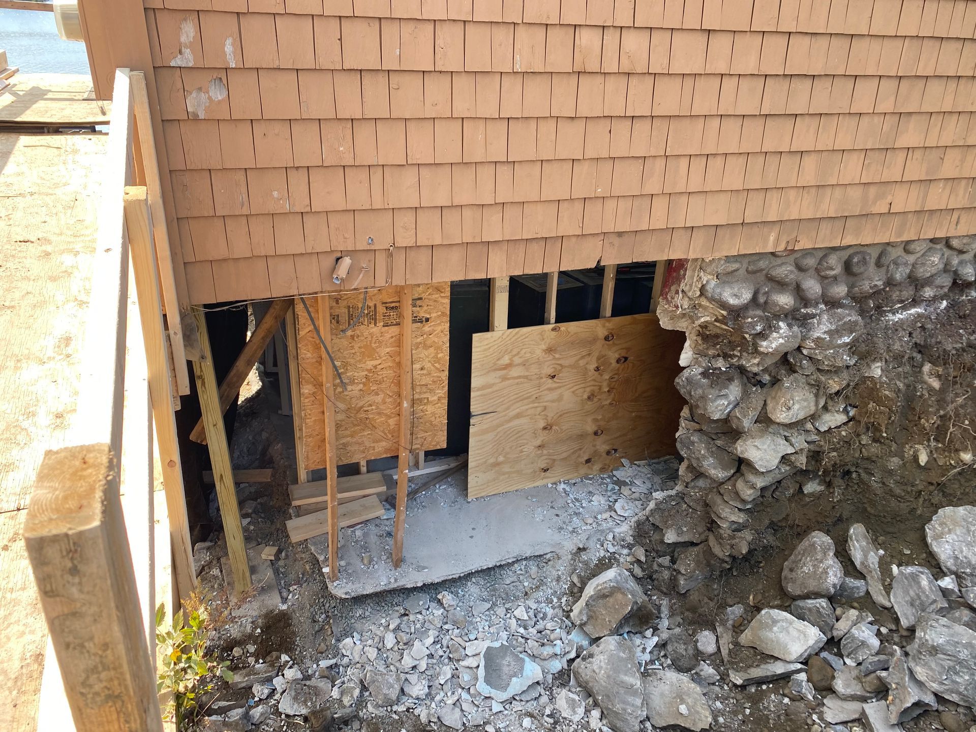 Construction site: exposed foundation and brick siding. Plywood and wooden supports visible near dirt and rocks.