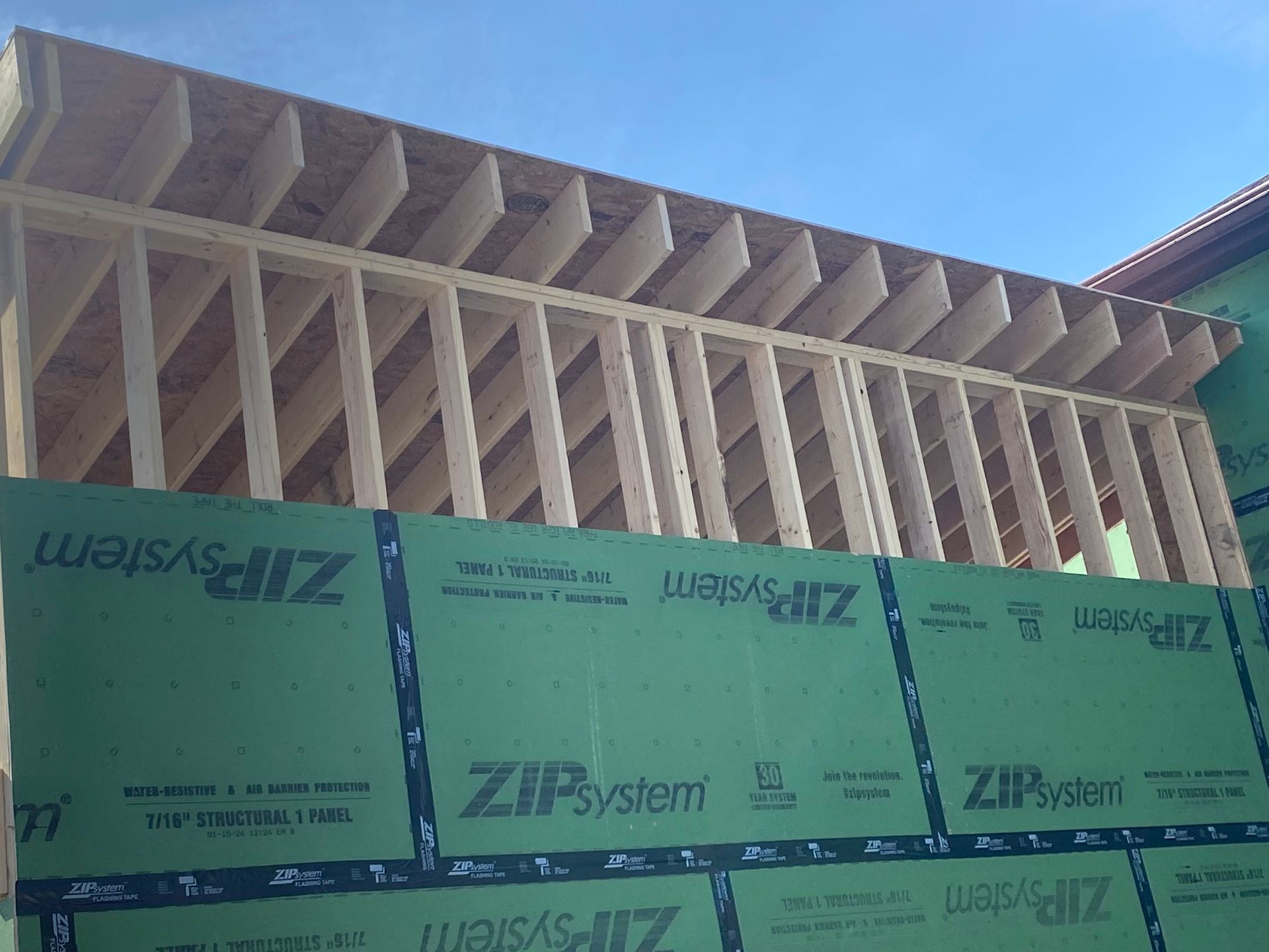 Construction of a building frame with wooden beams and green sheathing against a blue sky.