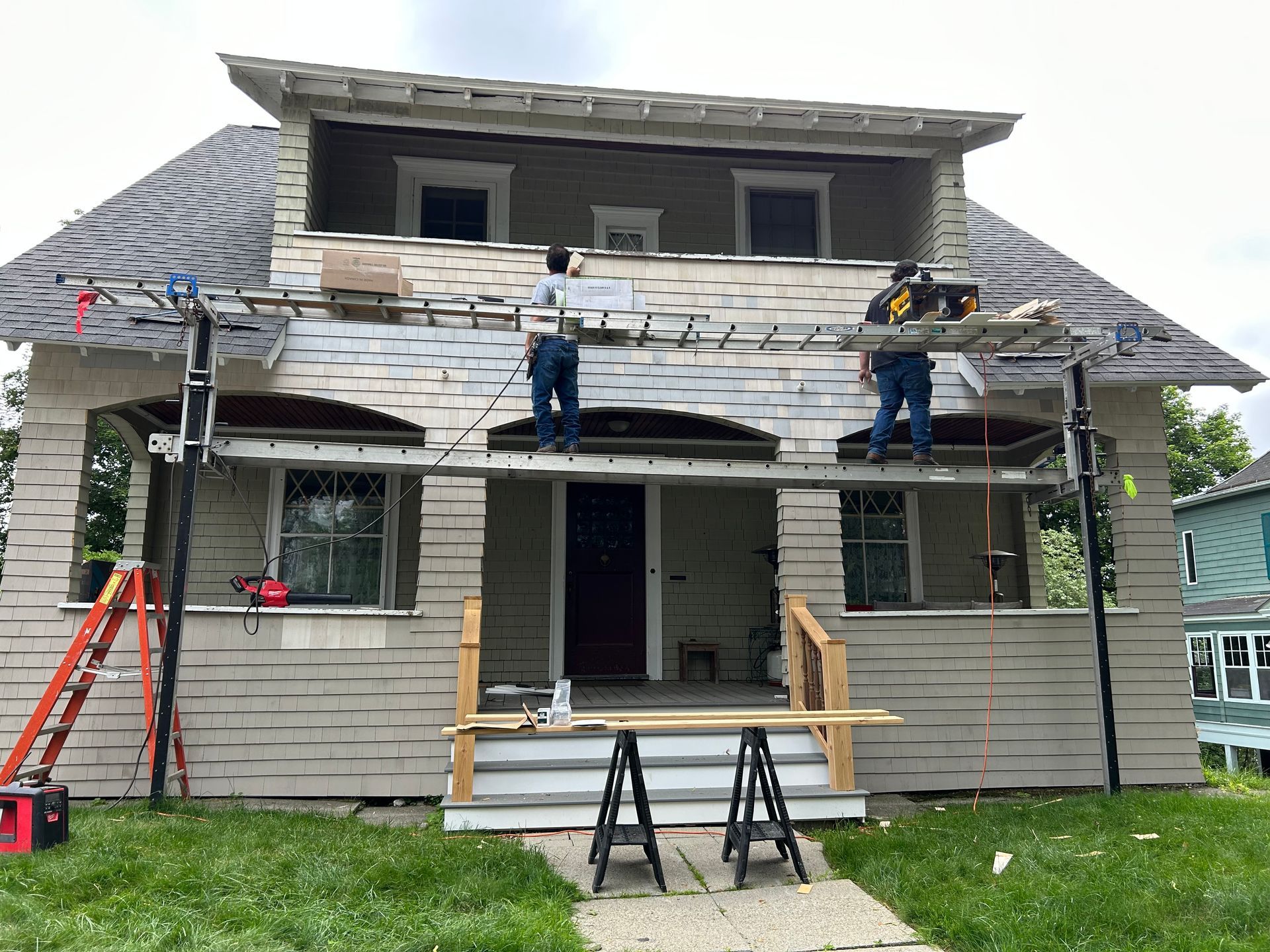 Two workers renovating a two-story house, using scaffolding. Gray siding, red door, cloudy sky.