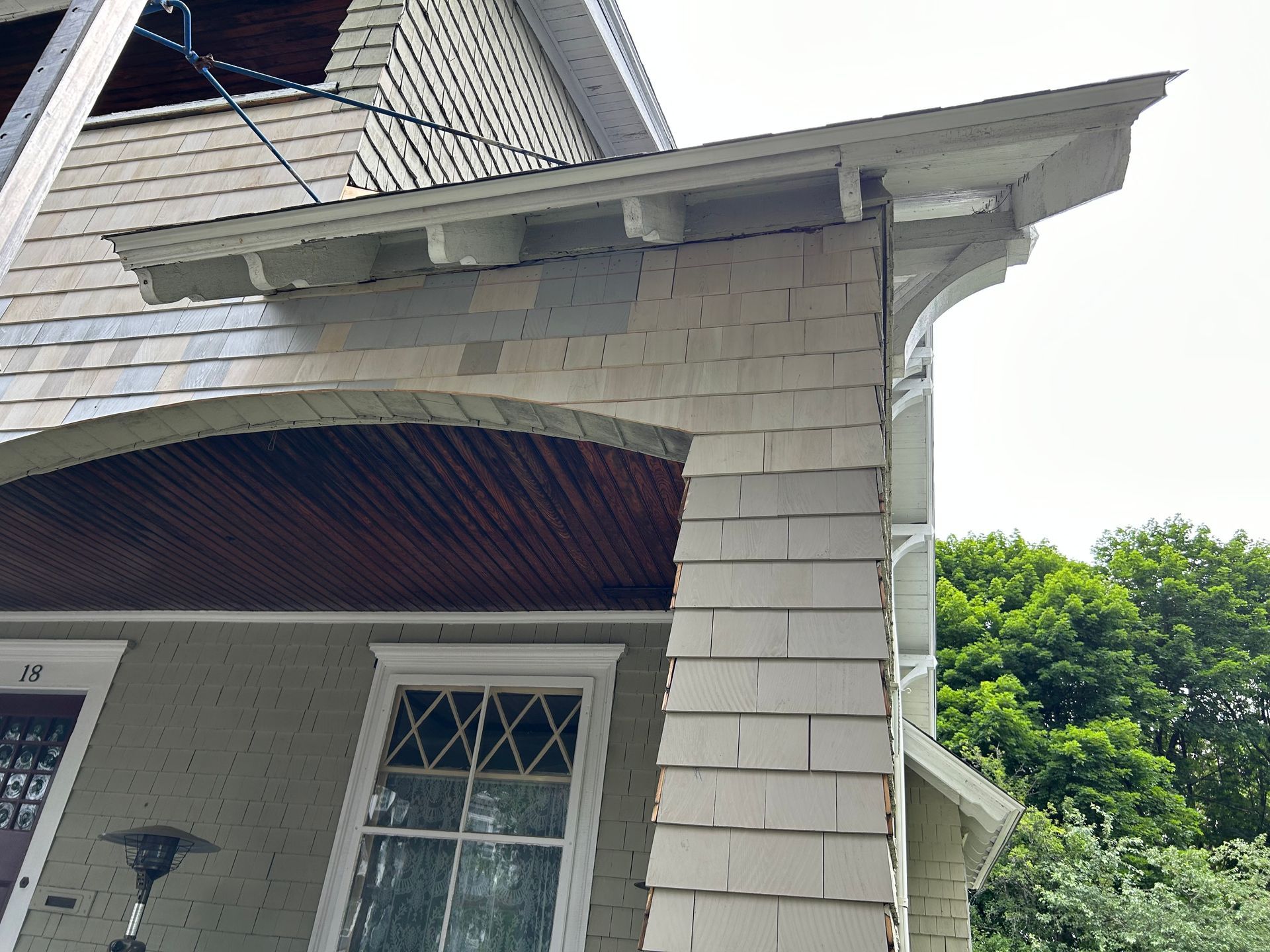 Exterior of a house with a porch. Shingled siding, arched entryway, white trim, and a decorative roof overhang.