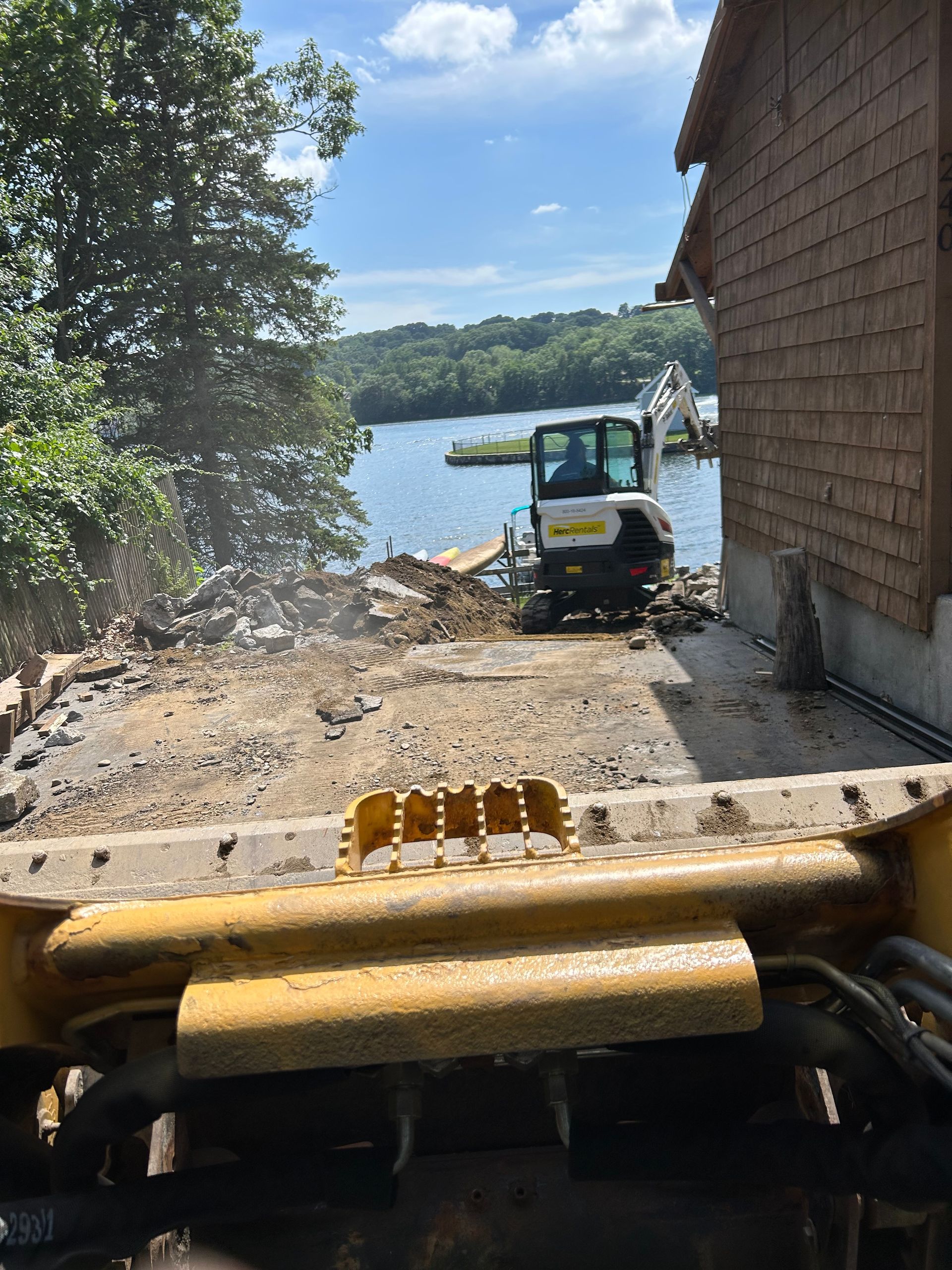 A construction site by water with a bobcat, a building, and trees on a sunny day.