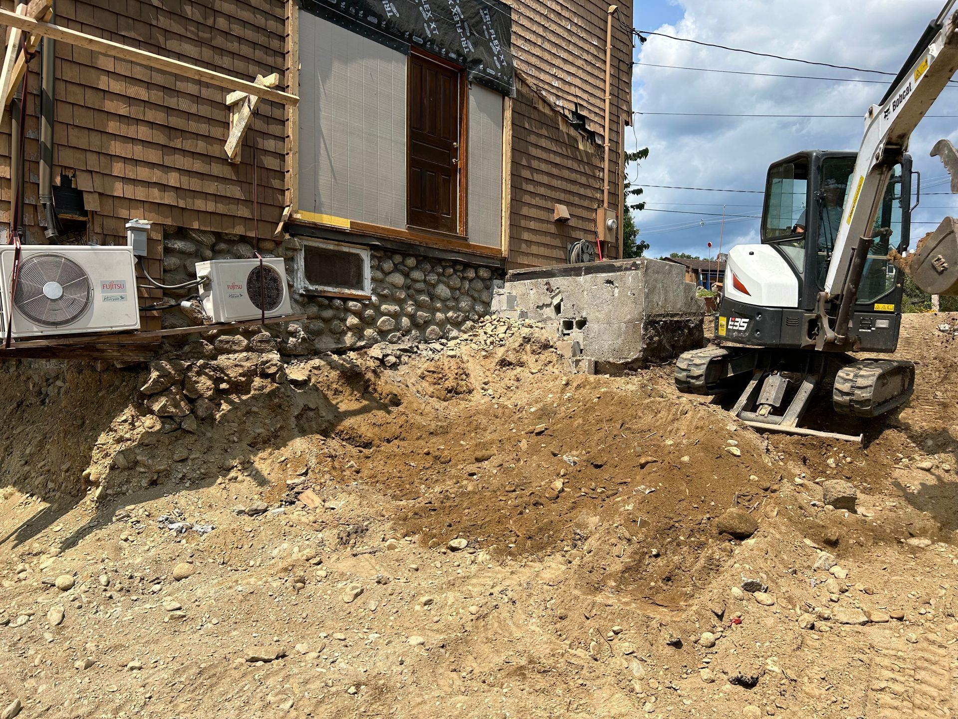 Excavator digging near a building with stone foundation and shingle siding; dirt mound in foreground.