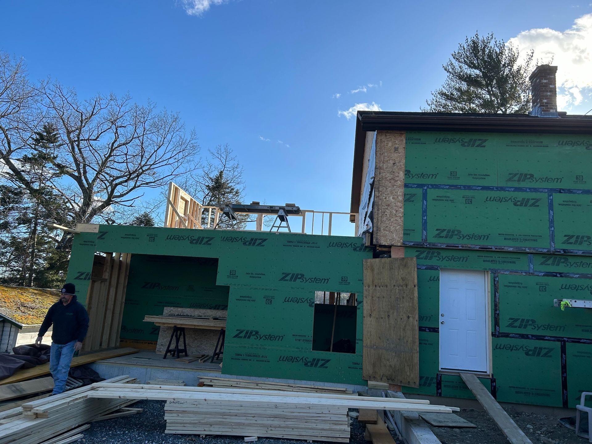 Construction site: a building with green sheathing. A person walks near lumber. Blue sky.