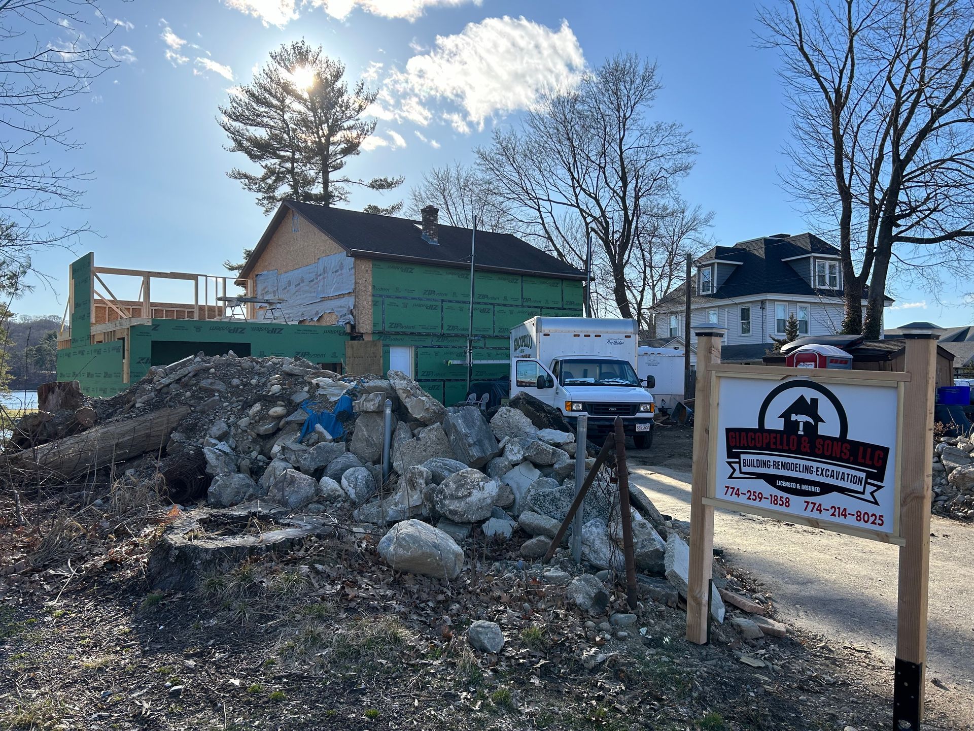 Construction site with building materials, a partially built structure, and a sign. Bright sunlight.
