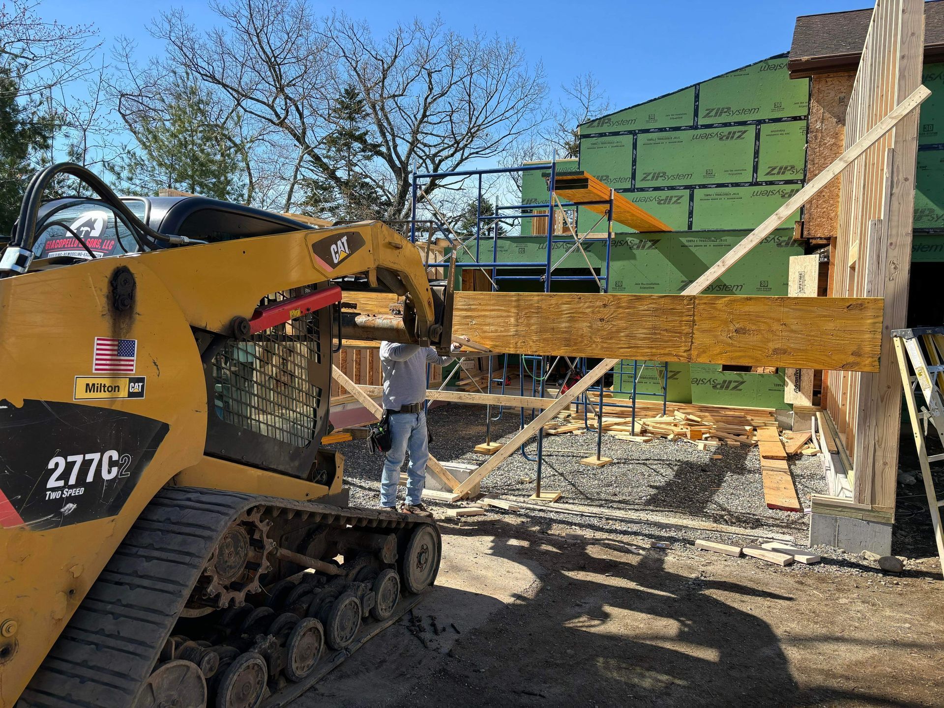 Yellow skid steer lifting a wooden beam at a construction site; a worker guides the beam.