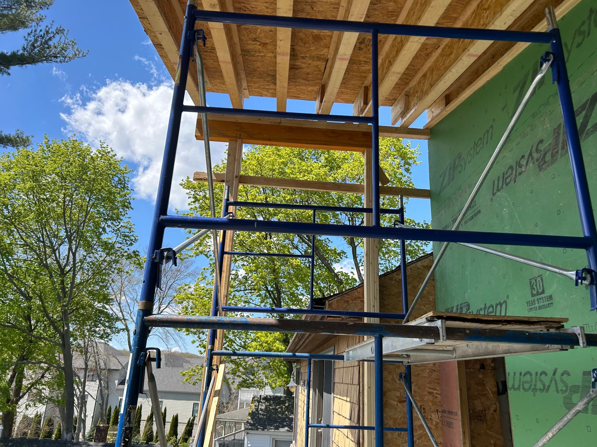 Blue scaffolding at a building construction site. The sky is blue with clouds, trees visible in the background.
