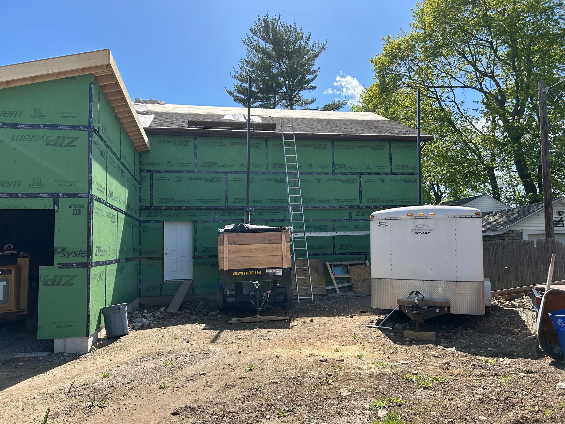 Construction site with green sheeting, a ladder, and a trailer on a dirt lot under a blue sky.