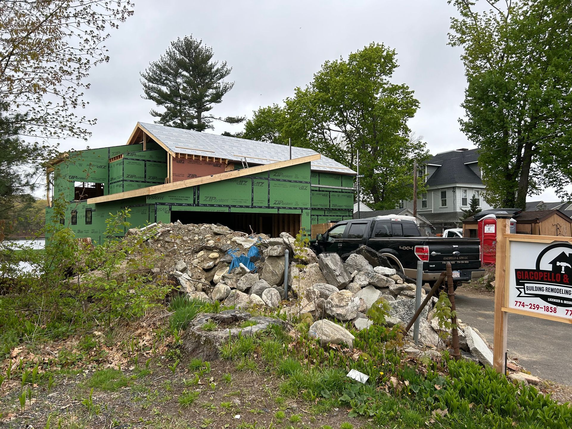 Construction site with partially built wooden building, green siding, rock pile, and a truck.