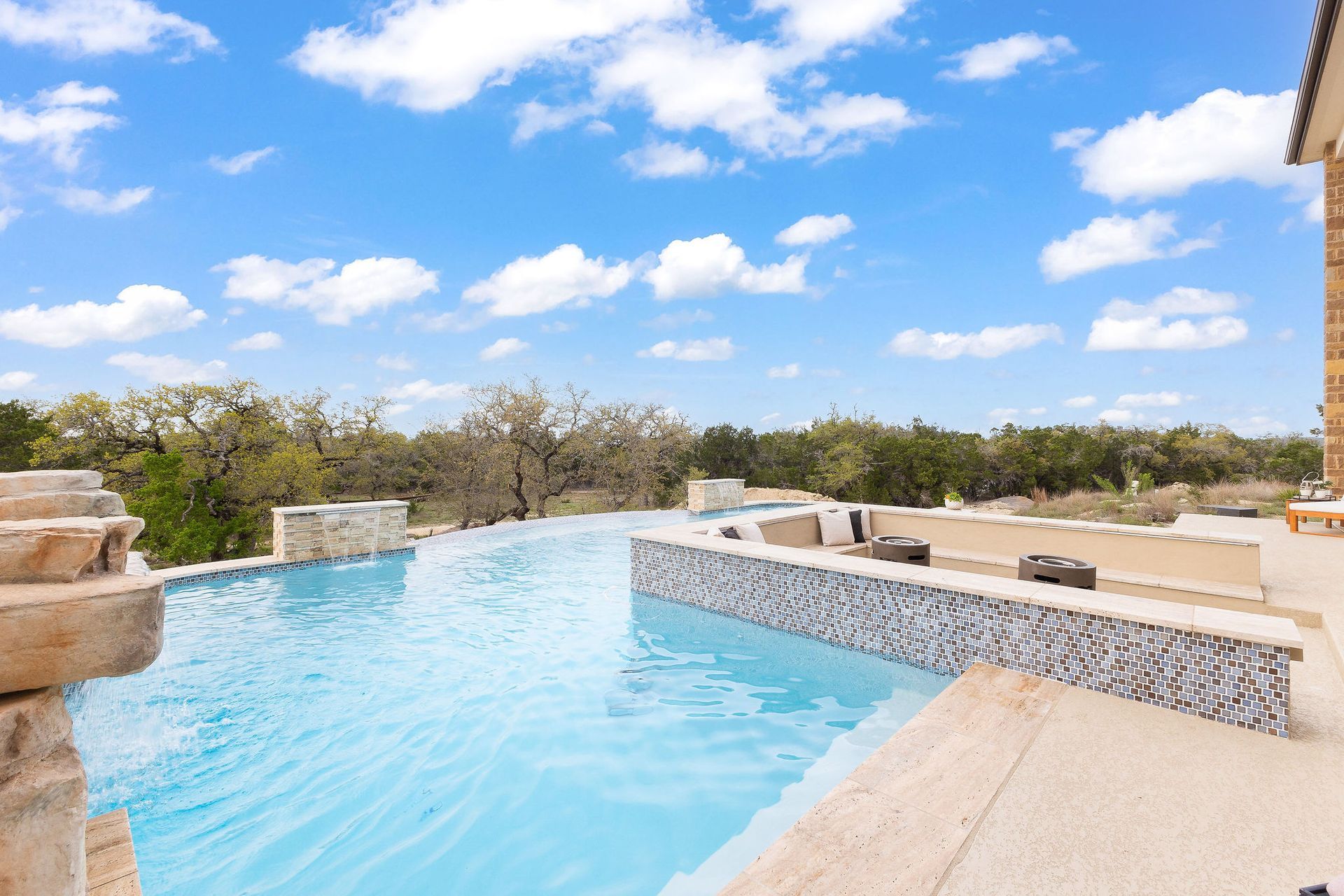 Infinity pool overlooking trees and blue sky, with tan stone deck and lounge chairs