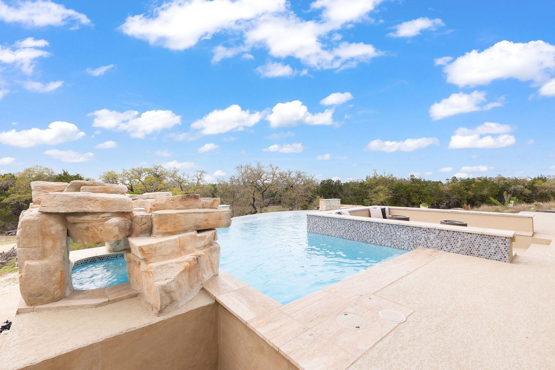 Stone hot tub beside a blue pool under a bright sky, with desert landscaping and lounge chairs.