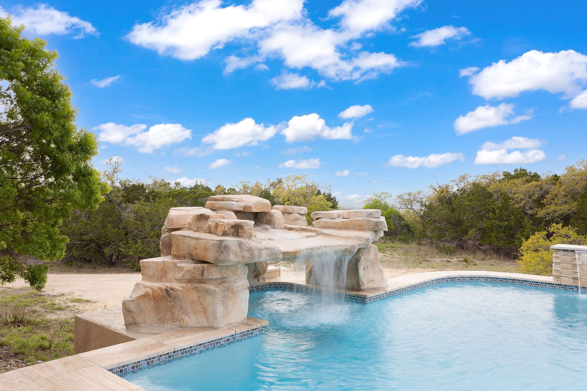 Stone waterfall spilling into a blue resort pool under a bright sky with trees around it