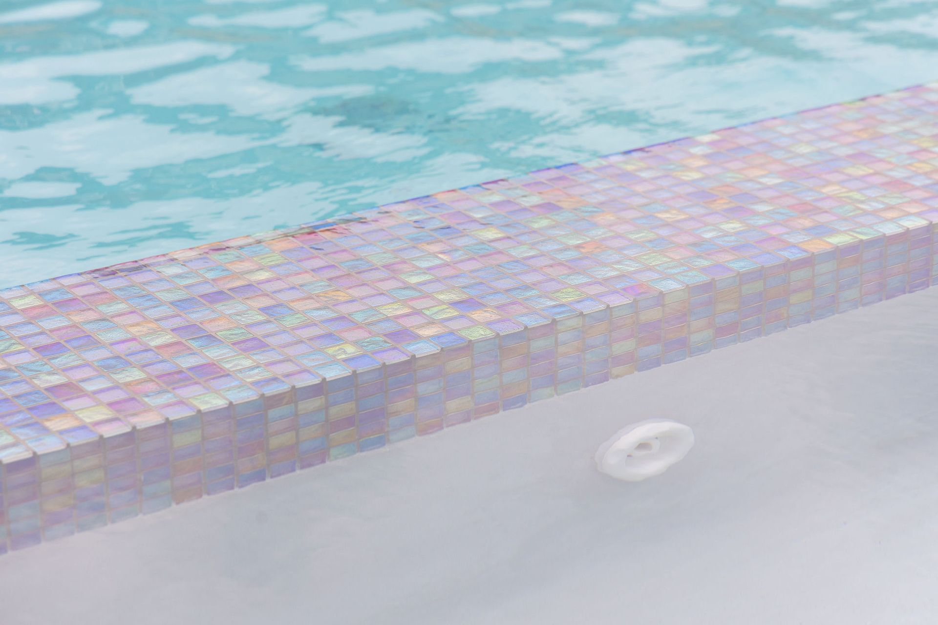 A tiled pool edge beside light blue water with a white drain cover in the foreground.