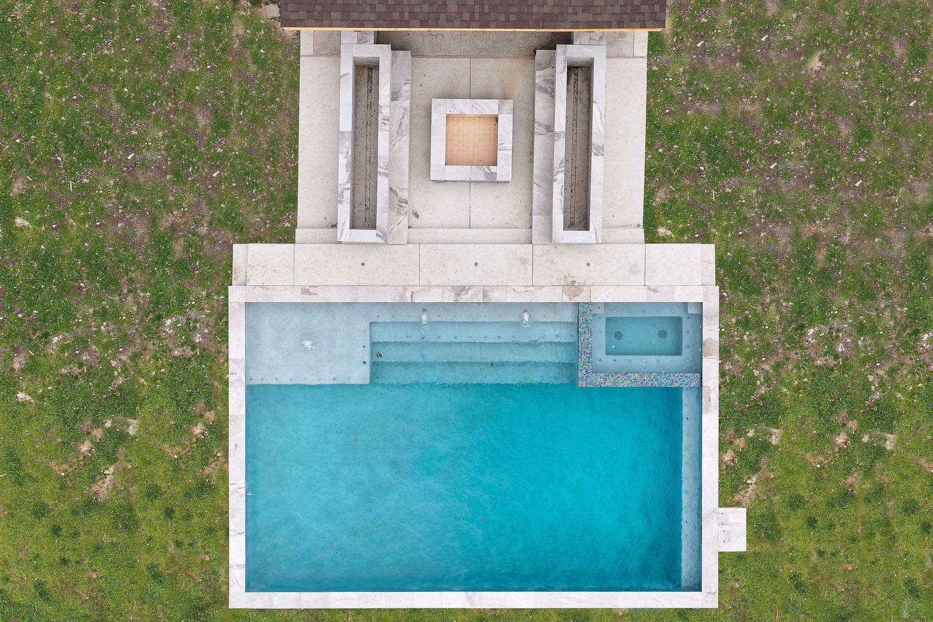 Aerial view of a rectangular turquoise pool beside a grassy yard and stone patio with loungers.