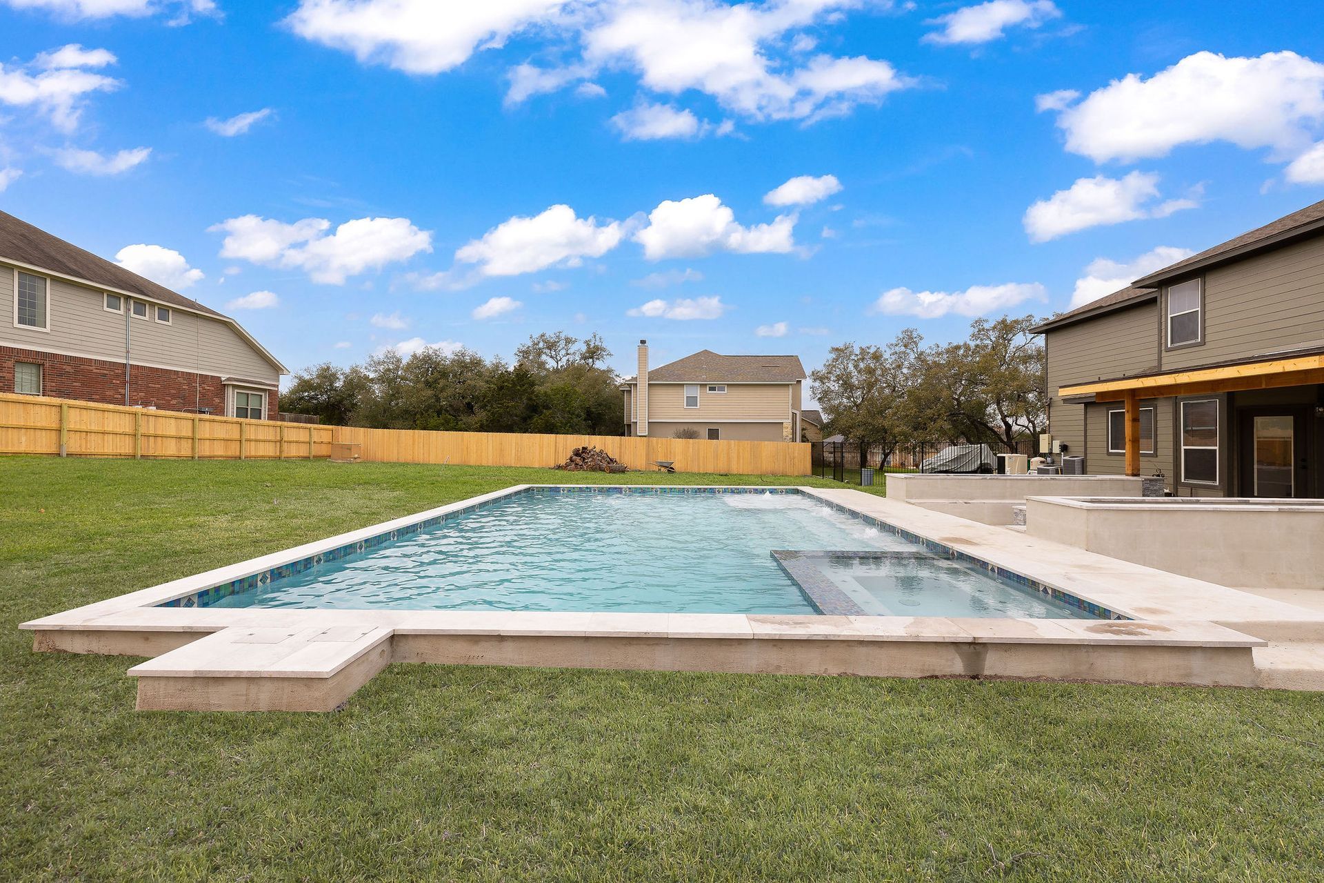 Rectangular backyard swimming pool with turquoise water, surrounded by grass and suburban houses under a blue sky