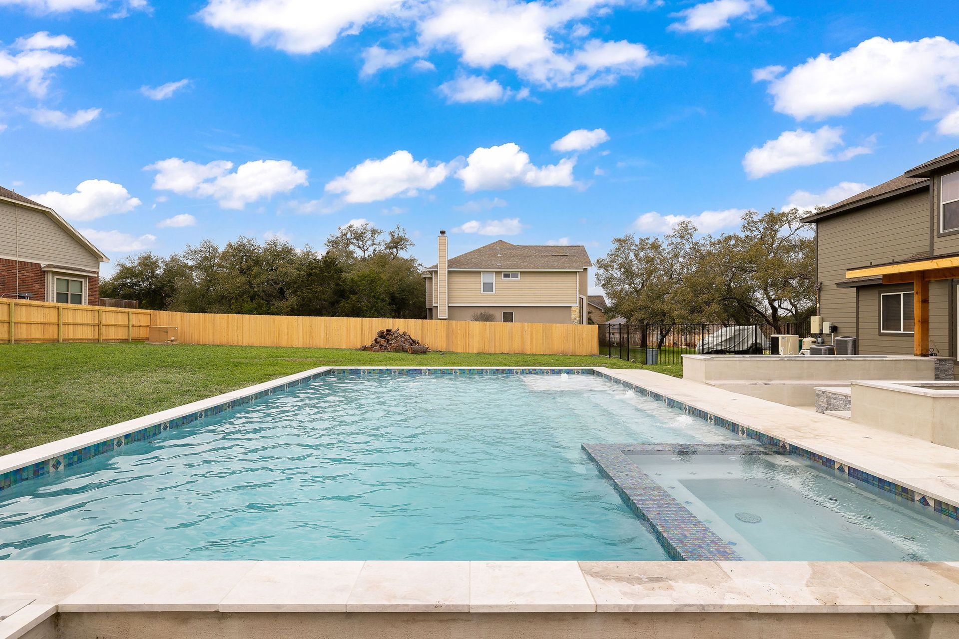 Backyard swimming pool with clear blue water, beige patio, grass, and houses under a partly cloudy sky