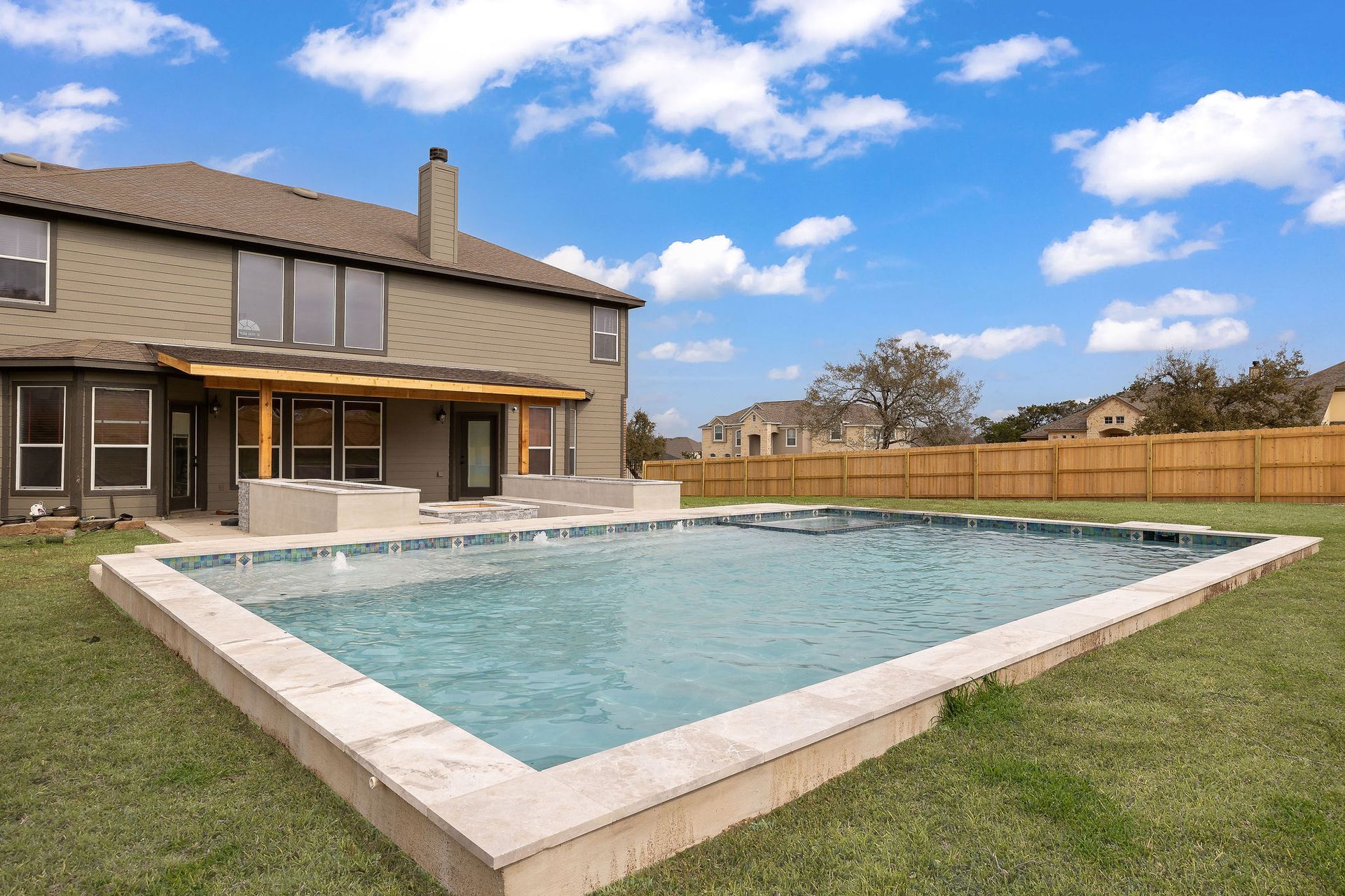 Backyard patio with a rectangular swimming pool beside a two-story house and wooden fence