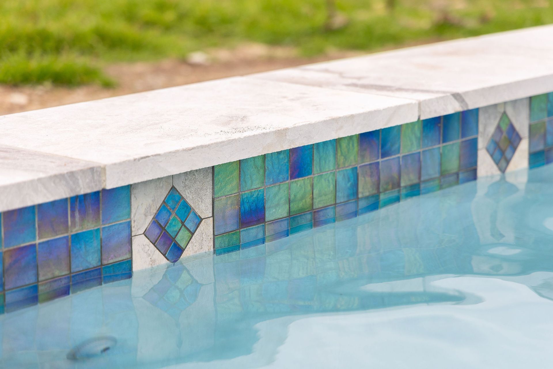 Blue-tiled swimming pool edge with white coping and clear water shimmering beside it
