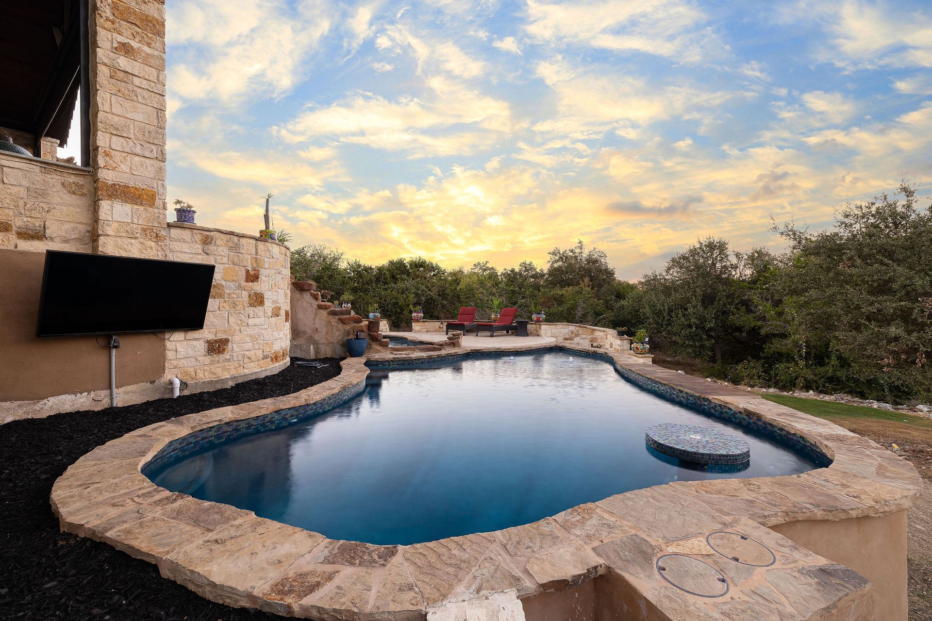 Stone patio with a curved backyard pool at sunset, bordered by trees and a stone house.