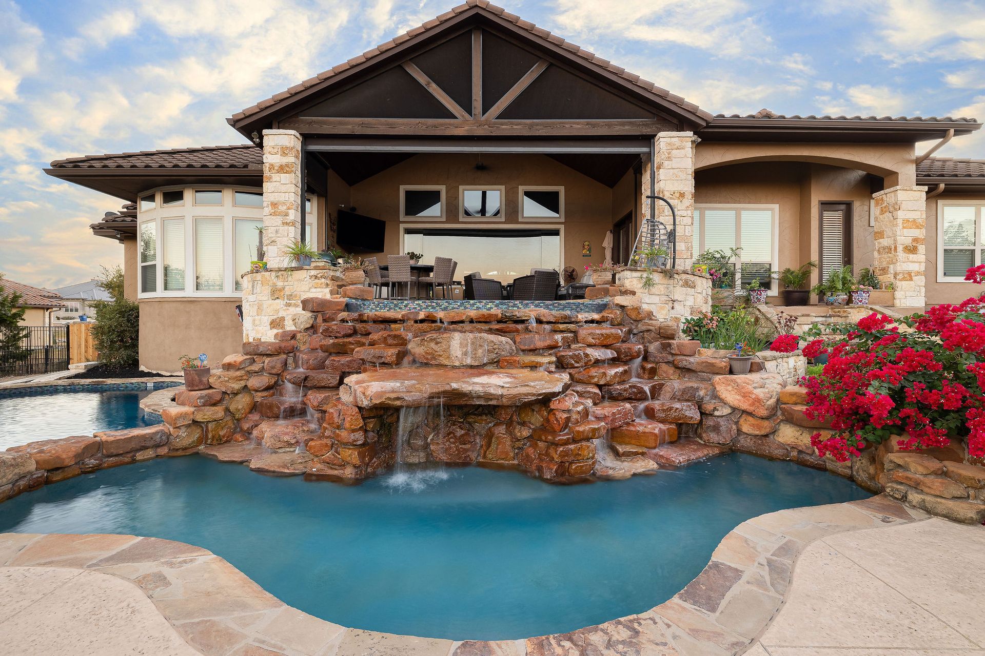 Luxury home patio with stone waterfall spilling into a blue pool, framed by flowers and stone columns