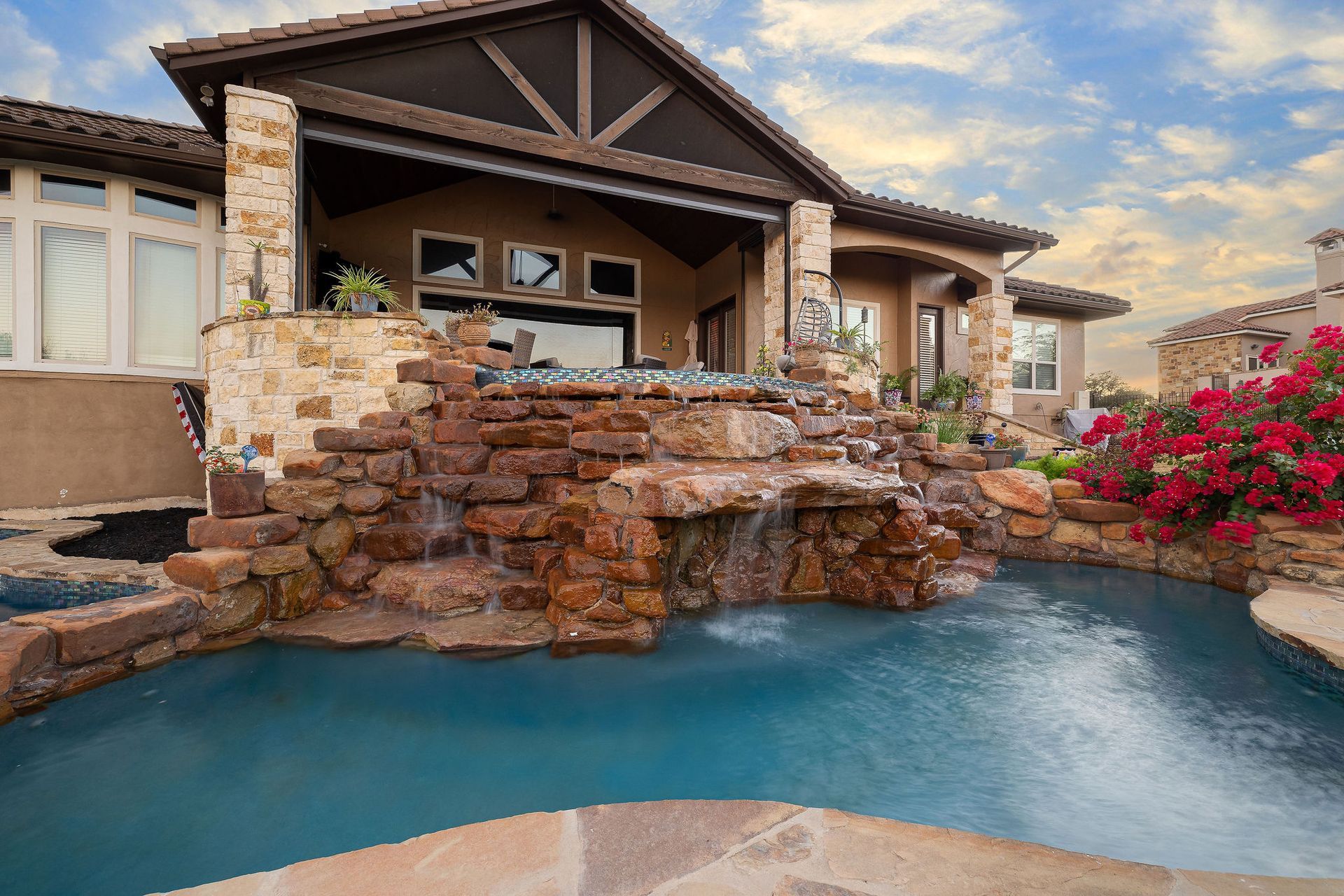 Backyard pool with rock waterfall and stone patio in front of a stucco house at sunset