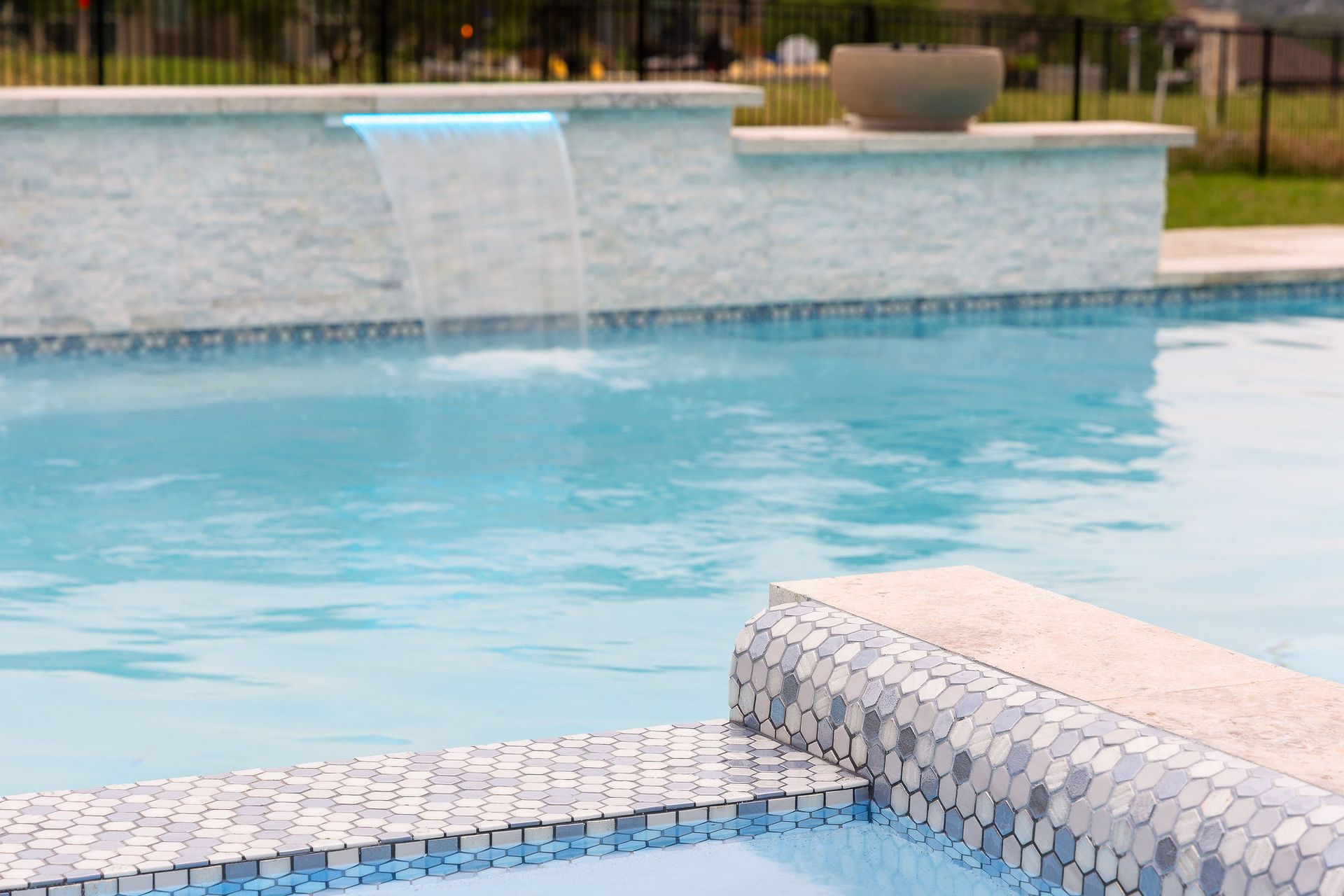 Outdoor swimming pool with stone patio, white lounge chairs, and trees under a partly cloudy blue sky