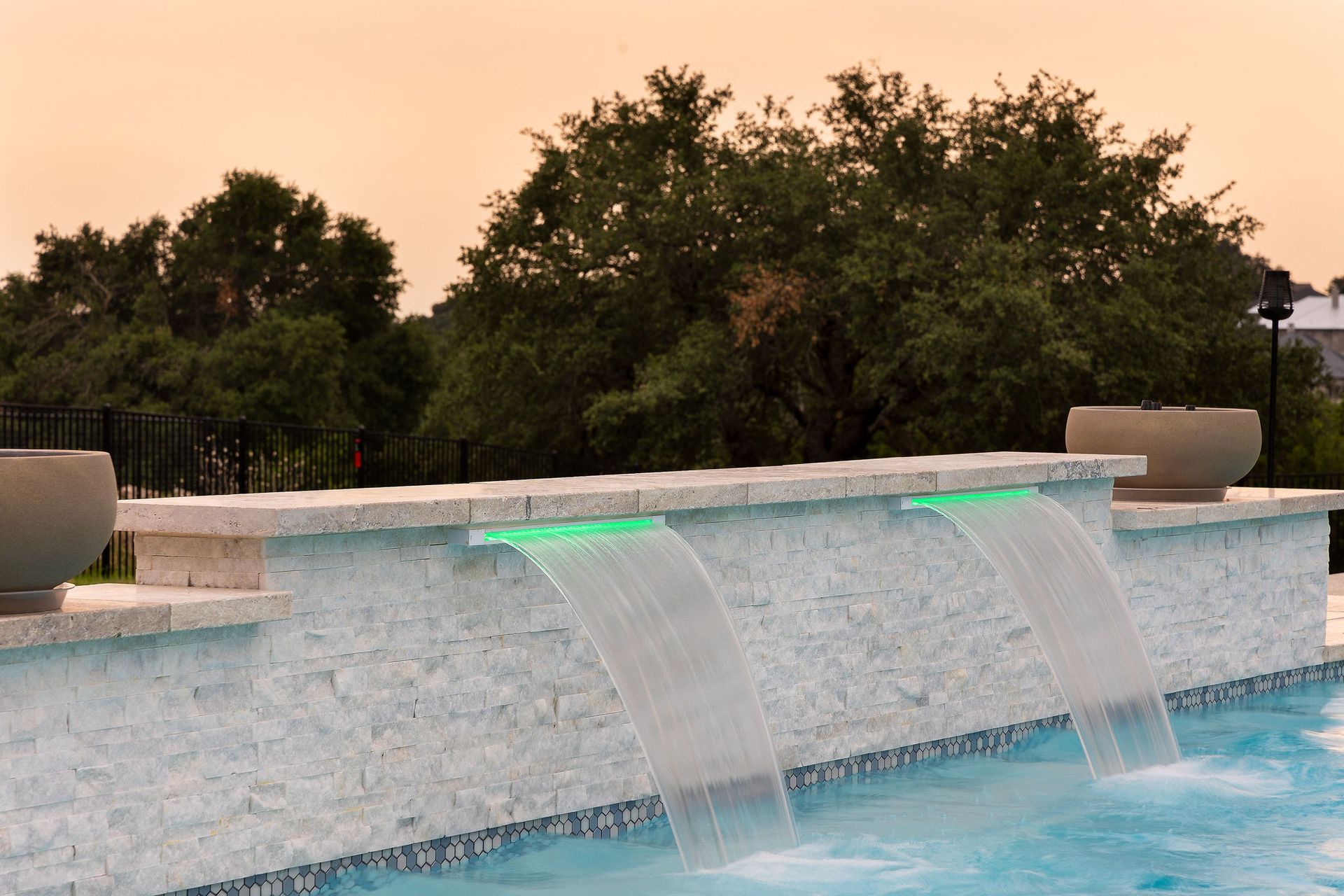 Modern pool waterfall with green LED lights at dusk and trees in the background