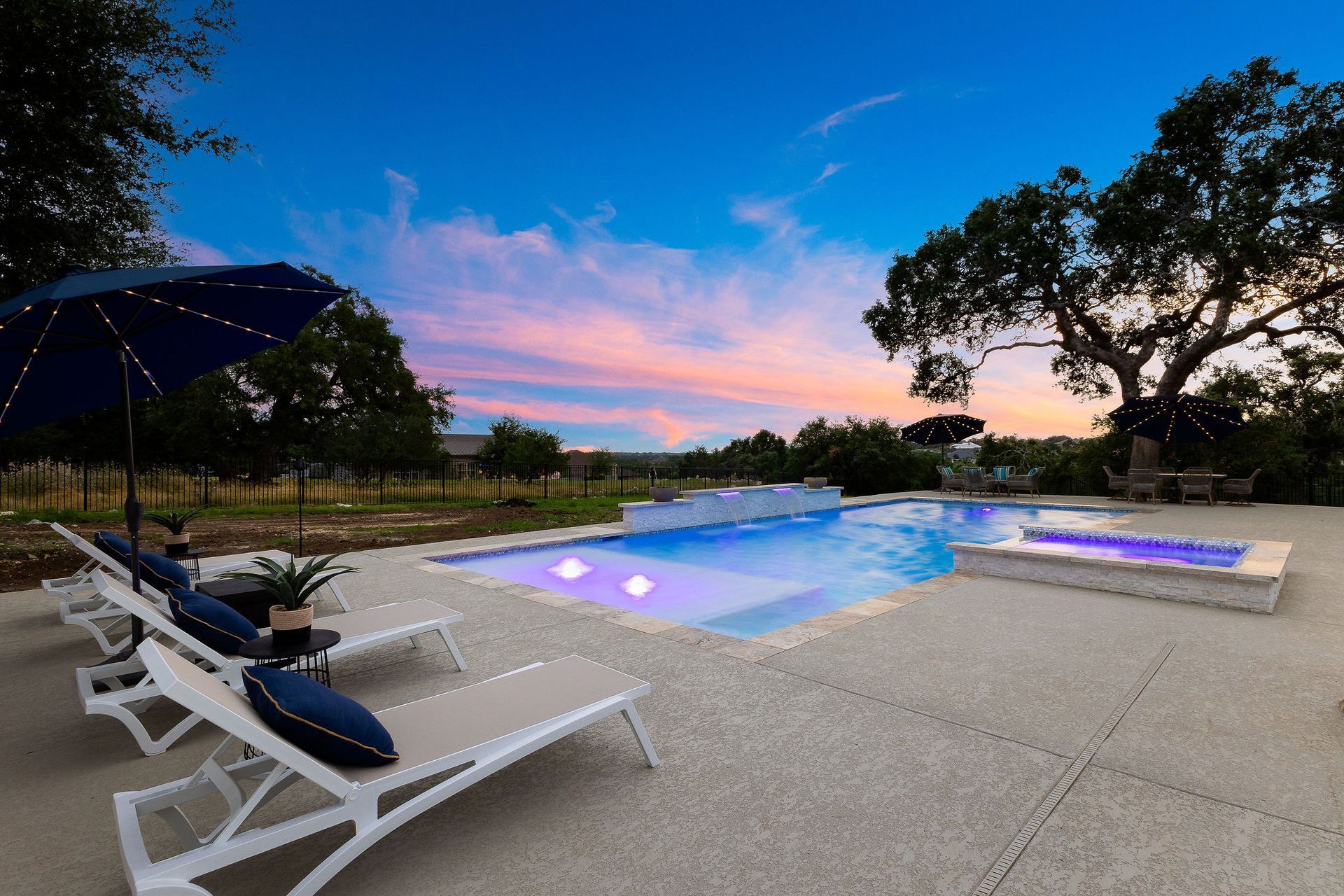 Backyard pool at dusk with glowing blue water, lounge chairs, umbrella, and trees against a pink sunset sky