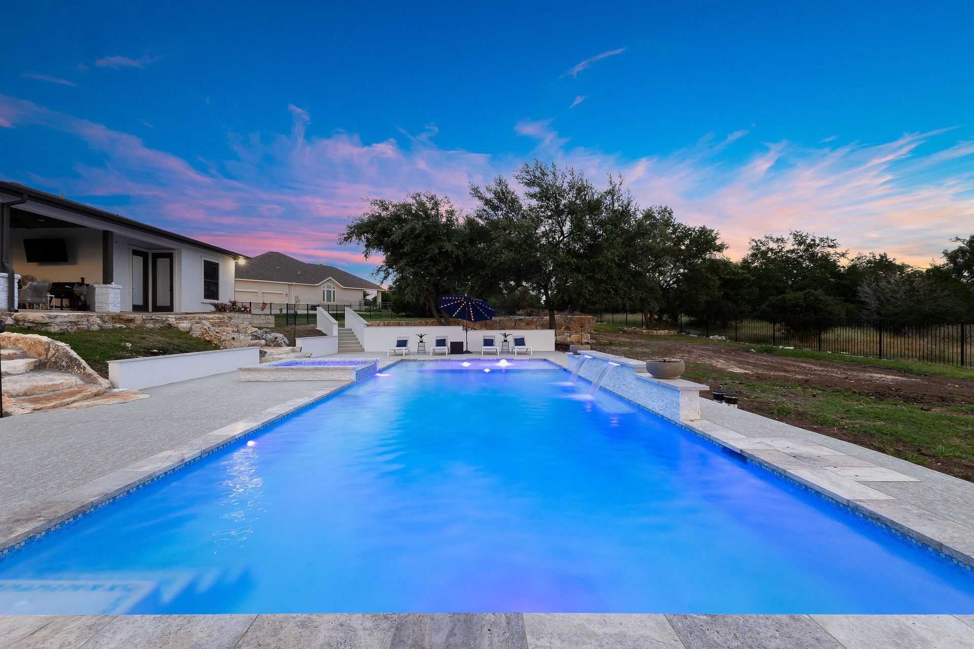 Backyard pool at sunset with blue water, stone deck, and a house beside trees and hills.