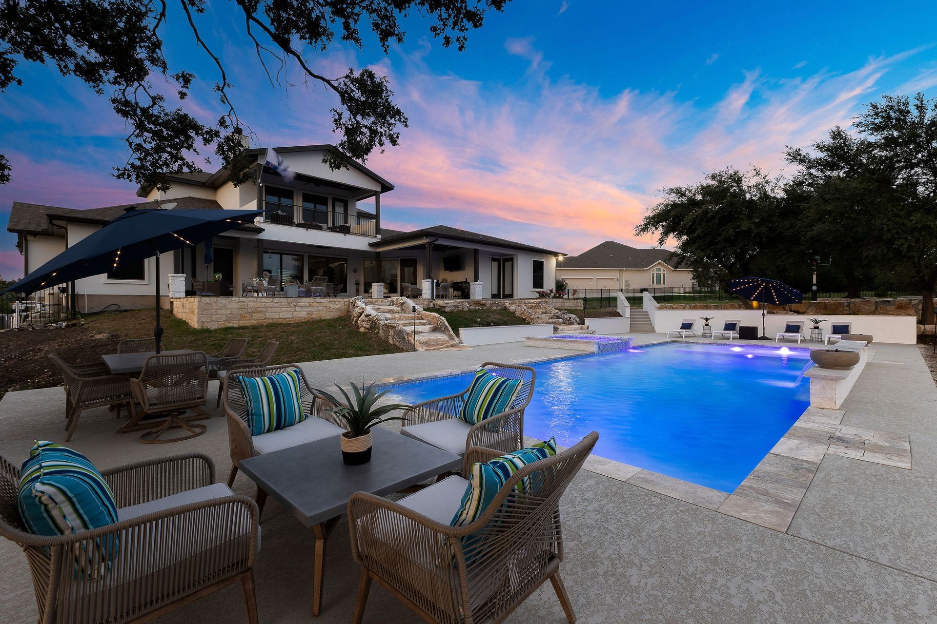 Backyard patio with a lit blue swimming pool, wicker chairs, and a modern house at sunset