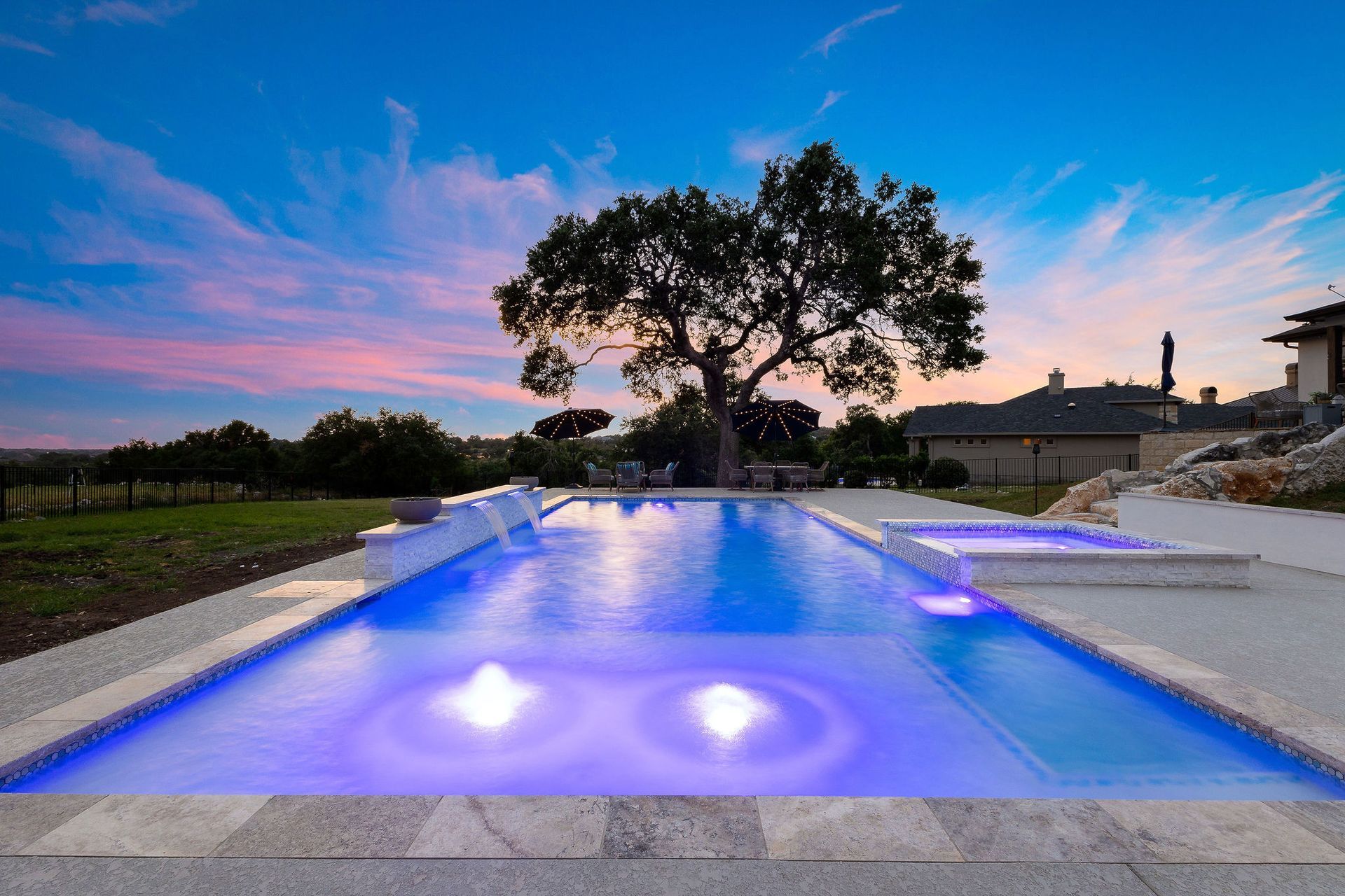 Modern backyard pool at dusk with blue-lit water, a large tree, and a colorful sunset sky