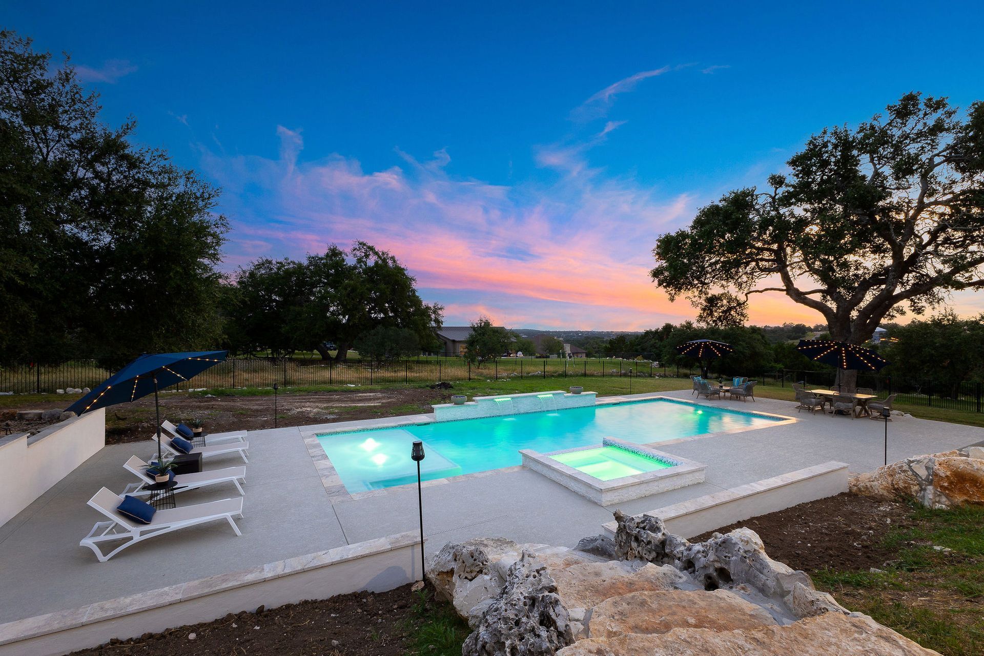 Backyard pool at dusk with lounge chairs, glowing blue water, and a pink-blue sky