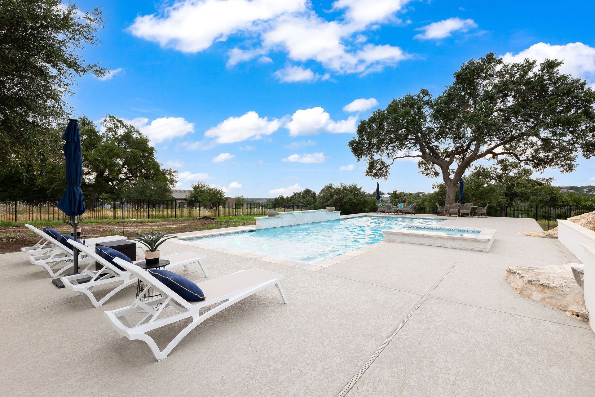 White lounge chairs beside a turquoise pool under a blue sky with trees and umbrellas