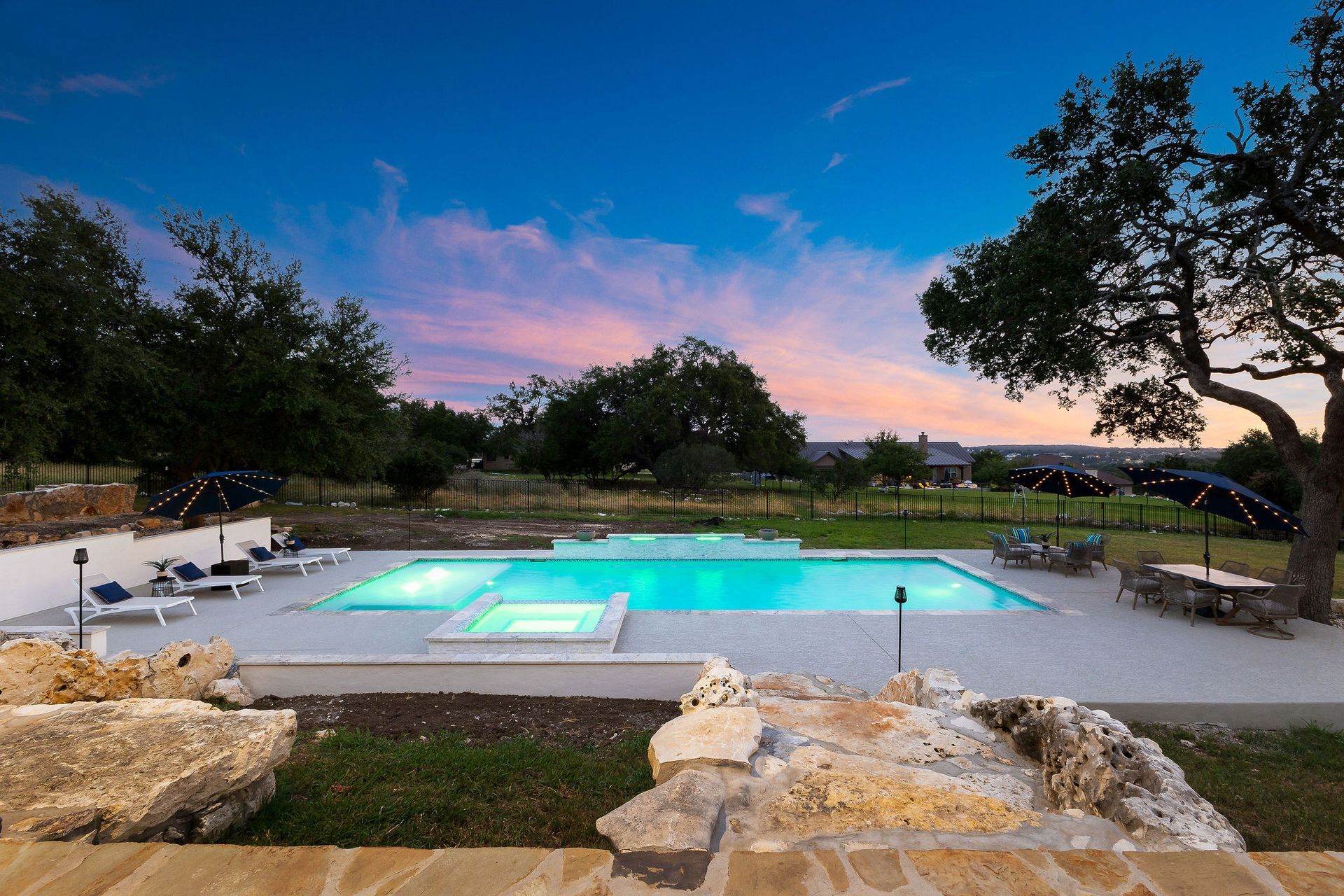 Backyard pool at dusk with turquoise water, stone patio, and trees under a pink-blue sky