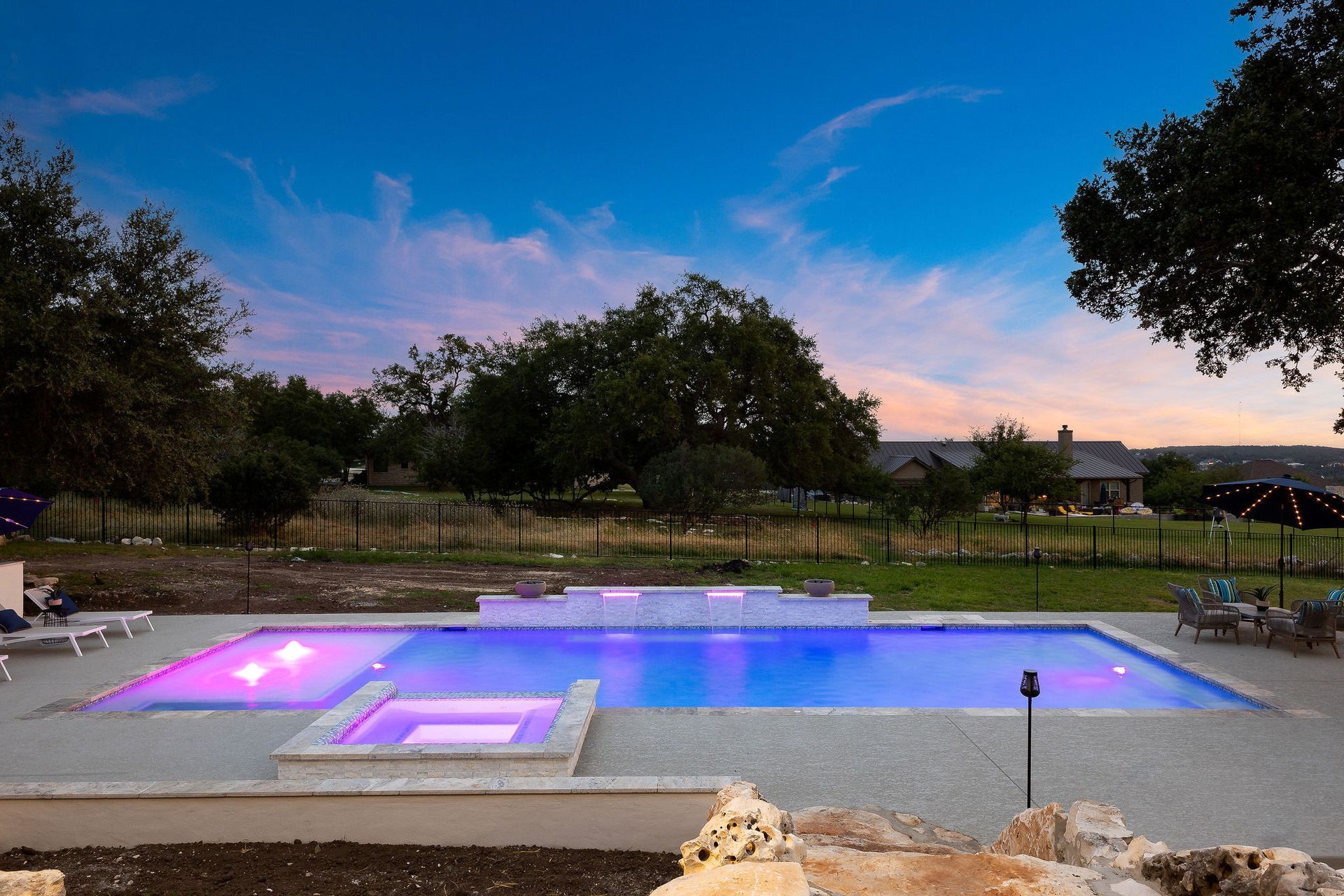 Backyard pool at dusk with purple lights, a deck, lawn, and trees under a twilight sky