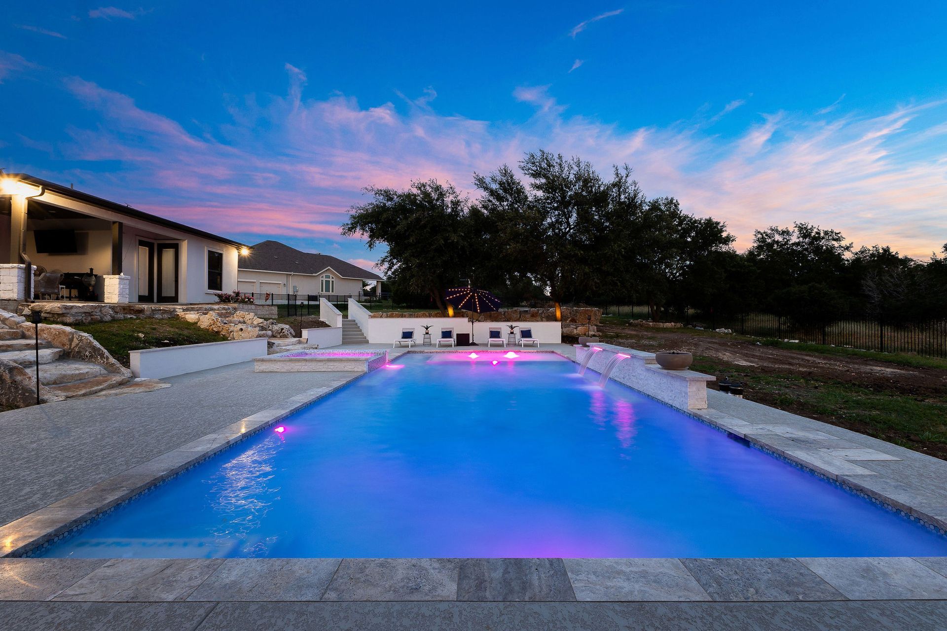 Modern backyard with a lit blue swimming pool at dusk beside a house and trees