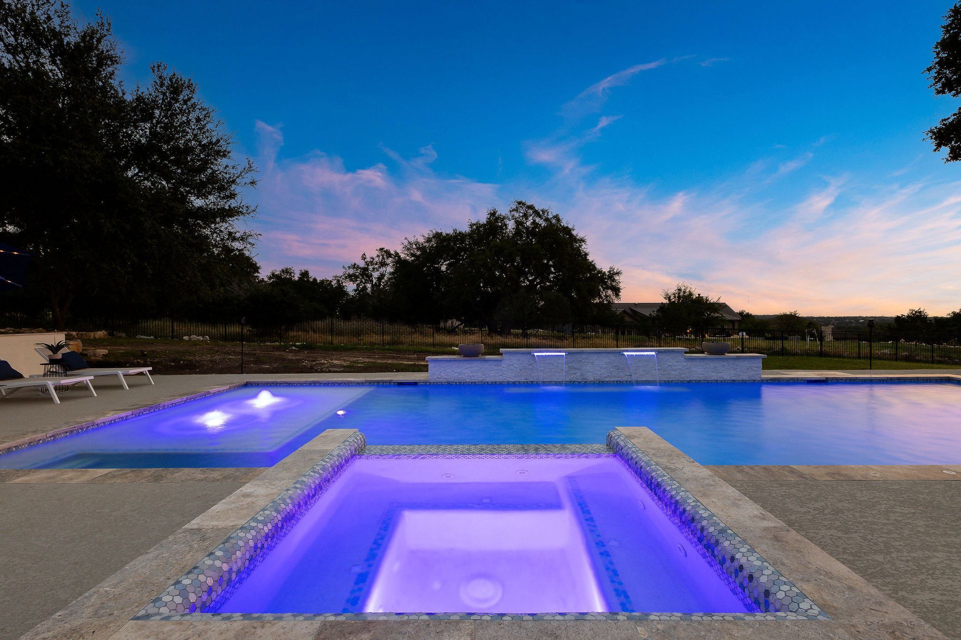 Lit backyard pool and hot tub glowing blue at dusk, with lounge chairs and trees nearby