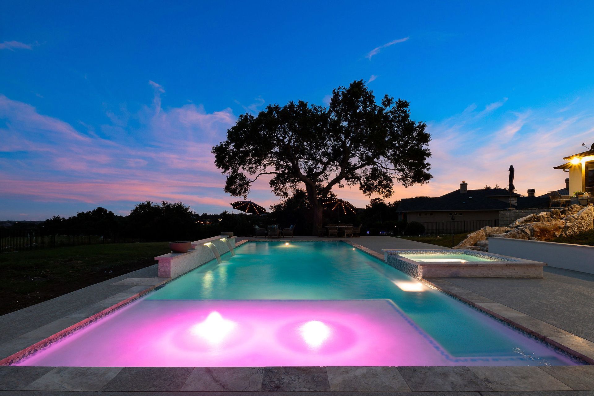 Backyard infinity pool at dusk with pink lights, reflecting a silhouetted tree and blue sky