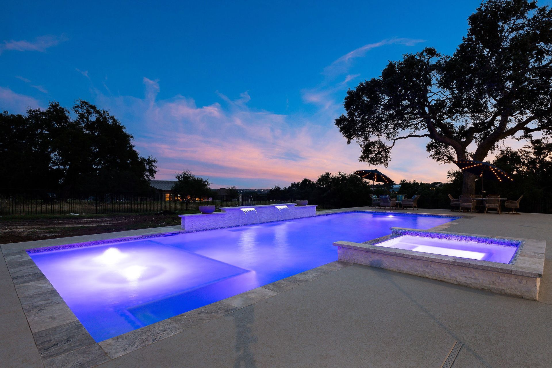 Modern backyard pool at dusk with glowing blue lights and trees silhouetted against a pink sky