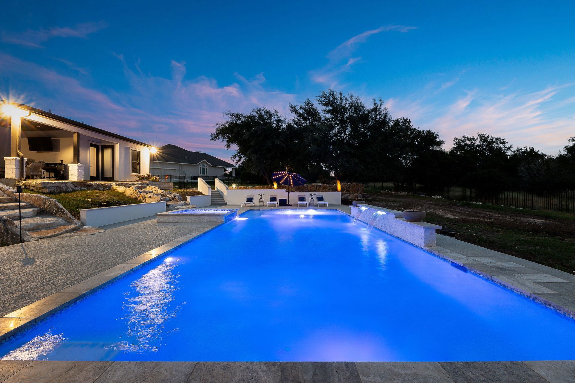 Illuminated backyard pool at dusk with glowing blue water beside a modern house