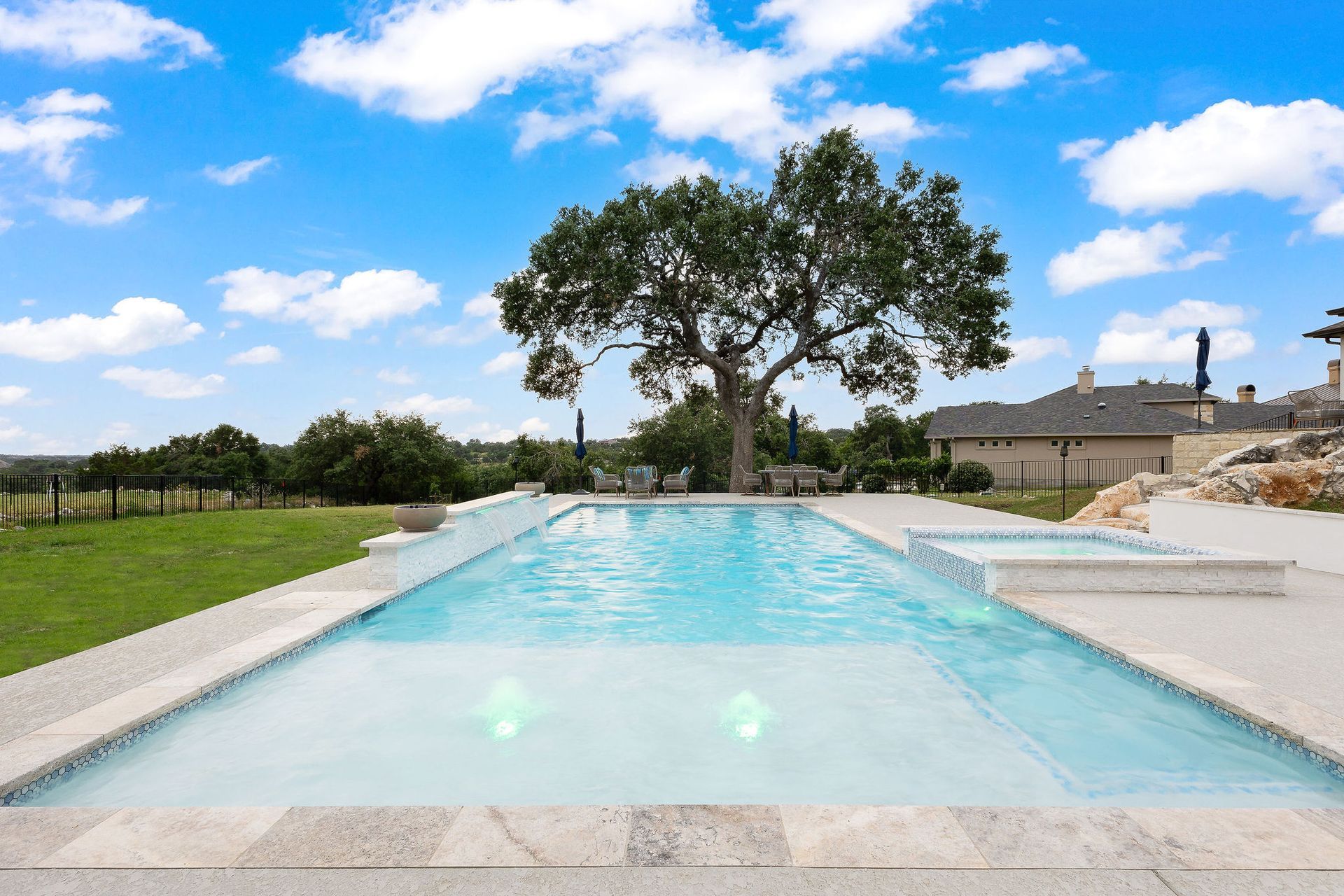 Infinity pool with clear blue water, a large tree, and a bright sky over a landscaped lawn and distant buildings