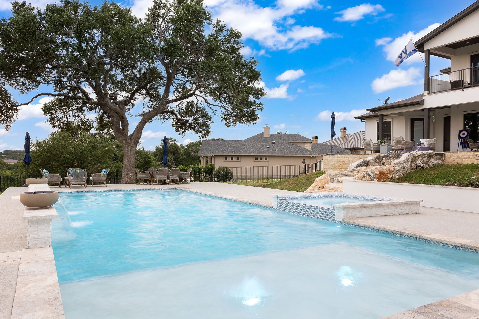 Backyard pool with blue water, stone patio, large tree, and a house under a bright sky