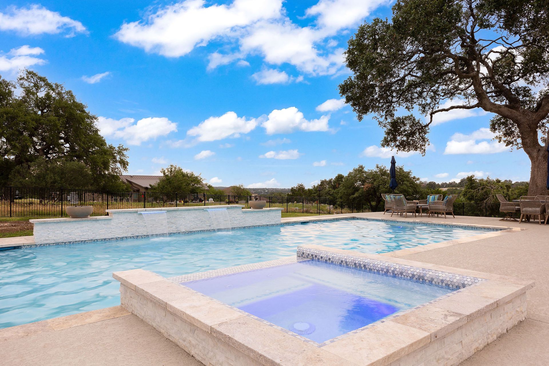 Stone resort pool and hot tub under a bright blue sky, with trees around the deck.