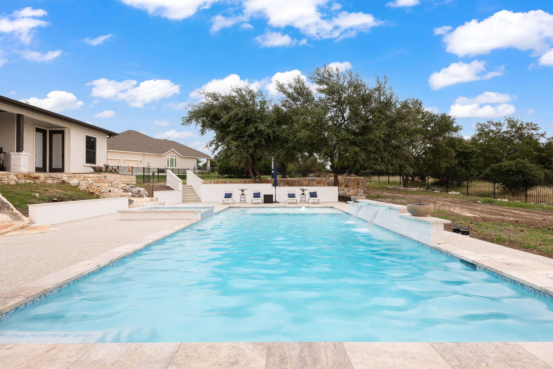 Outdoor swimming pool with clear blue water beside a modern house and trees under a partly cloudy sky