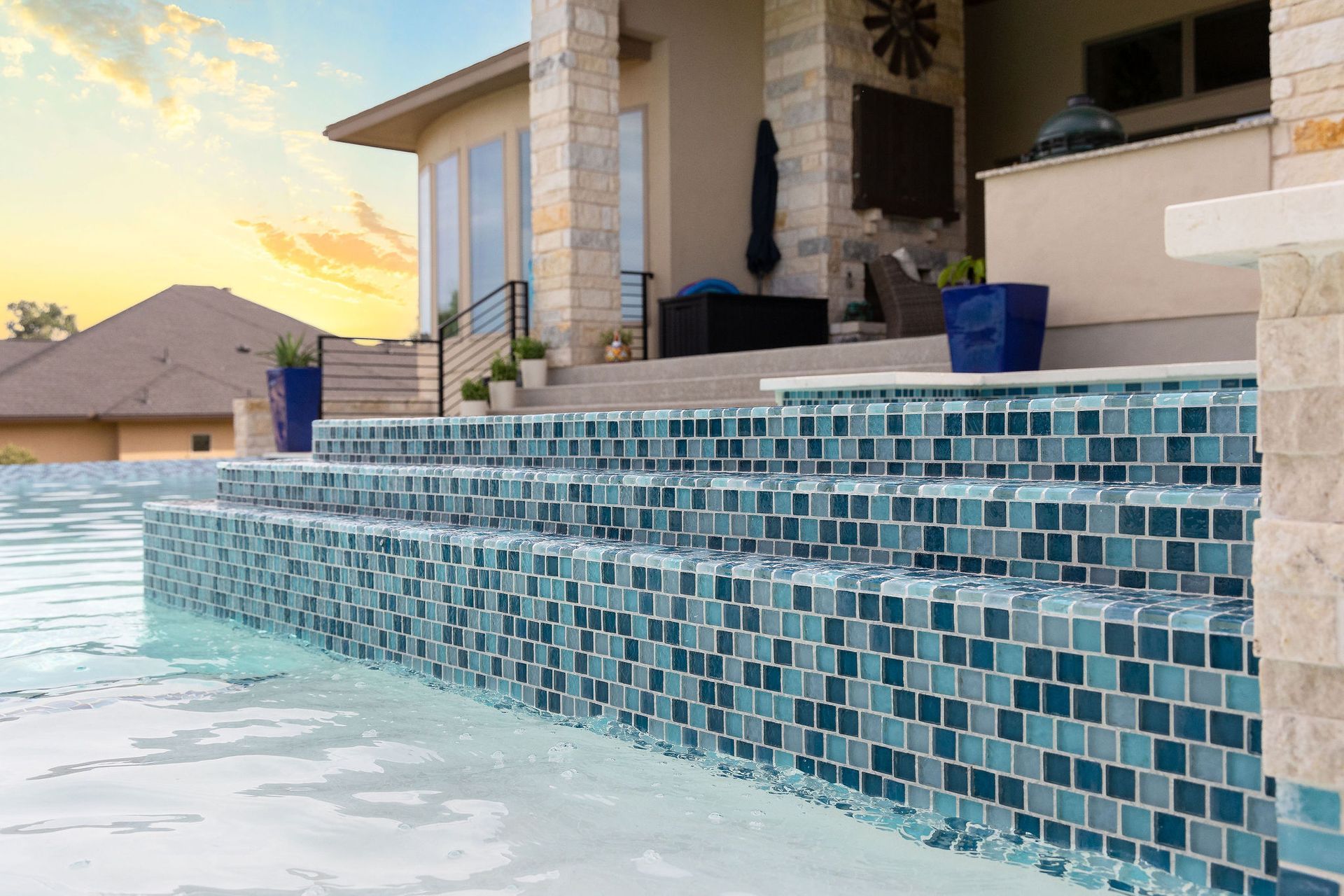 Backyard pool with blue mosaic tile steps and a stone patio at sunset
