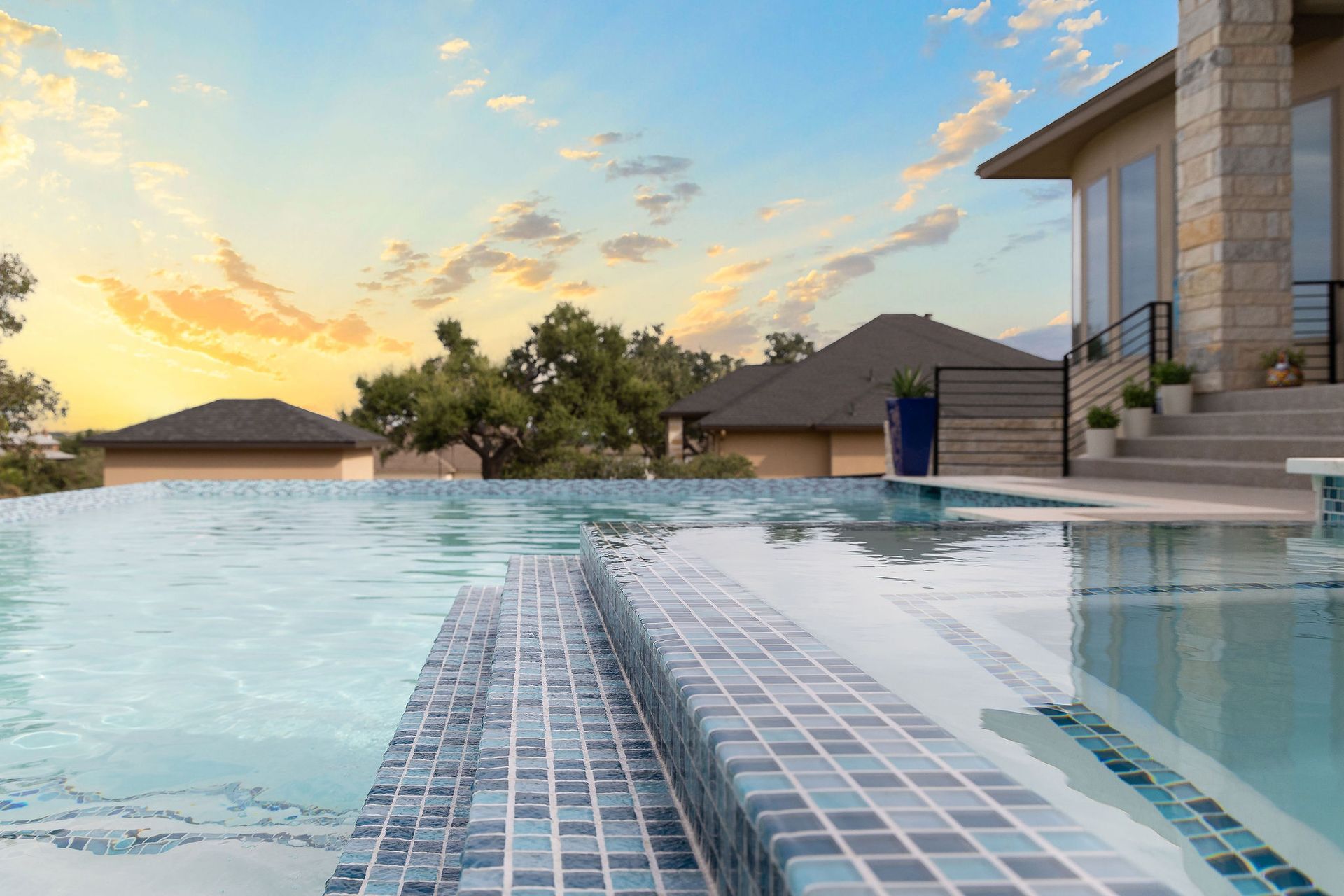 Modern infinity pool with tiled steps, patio, and sunset sky over distant hills
